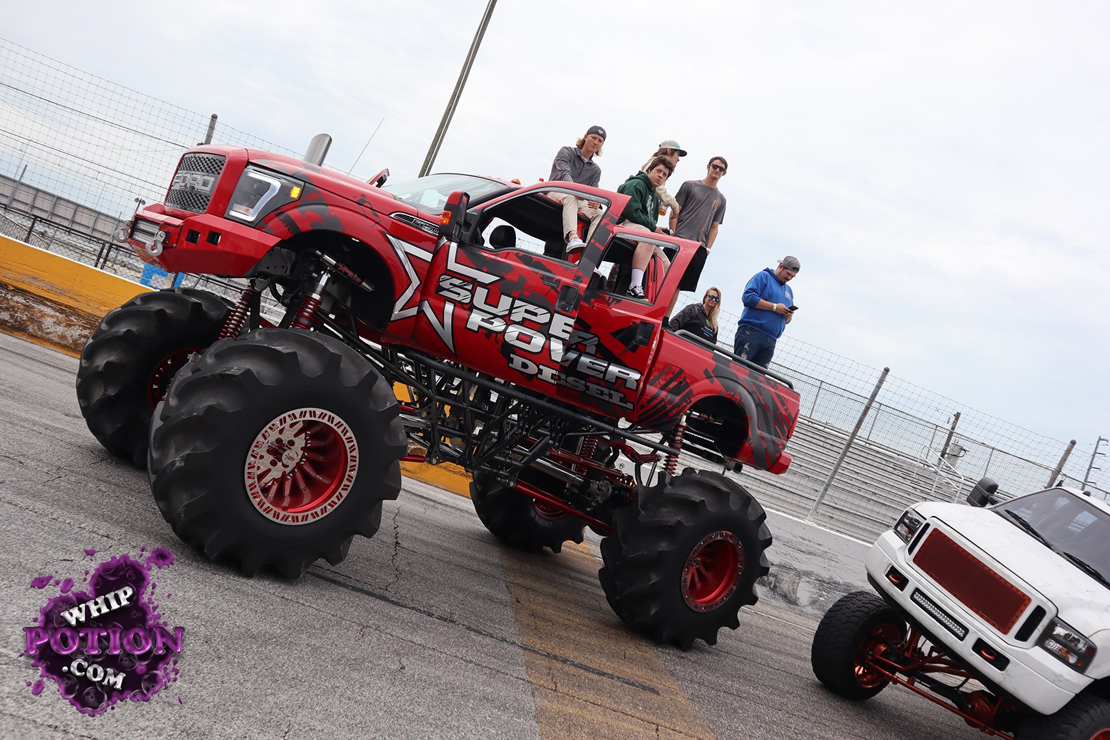 A bright red lifted pickup dominates the frame, riding on massive deep-tread tires with red-and-white wheels and exposed suspension. Bold black graphics and a large “Super Power Diesel” side decal give it an aggressive custom look, while several people sit and stand on the truck for scale. Shot at a racetrack under an overcast sky, the scene feels lively and oversized.