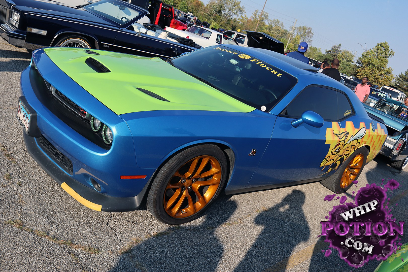 A Dodge Challenger gets a wild custom treatment here, with a bright blue body, neon green hood, and bronze wheels setting off the whole package. The cartoon-style side graphics and matching windshield banner give it a playful street-machine vibe, while the clean stance and tinted glass keep it looking sharp and purposeful.