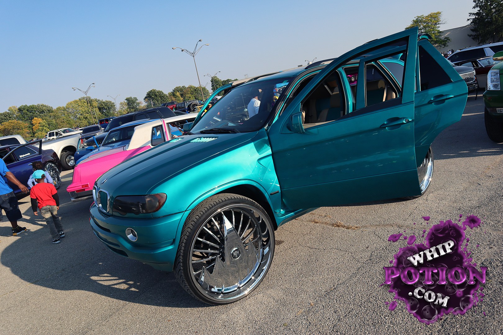 A teal BMW X5 gets the custom treatment with oversized chrome wheels, a slammed-looking stance, and both passenger doors swung wide to show off the clean tan interior. The metallic paint pops hard against the body’s chunky SUV lines, and the big-lip rims give it that full show-ready, attention-to-detail feel.
