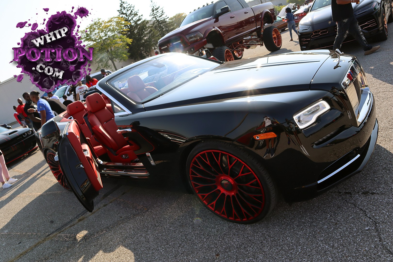 A black Rolls-Royce Wraith sits on deep red and black custom wheels, with the coach doors swung open to a vivid red leather cabin. The glossy paint, tall grille, and flying lady hood ornament give it that unmistakable luxury presence, while the color-matched interior and bold wheel choice add a modern show-car edge.