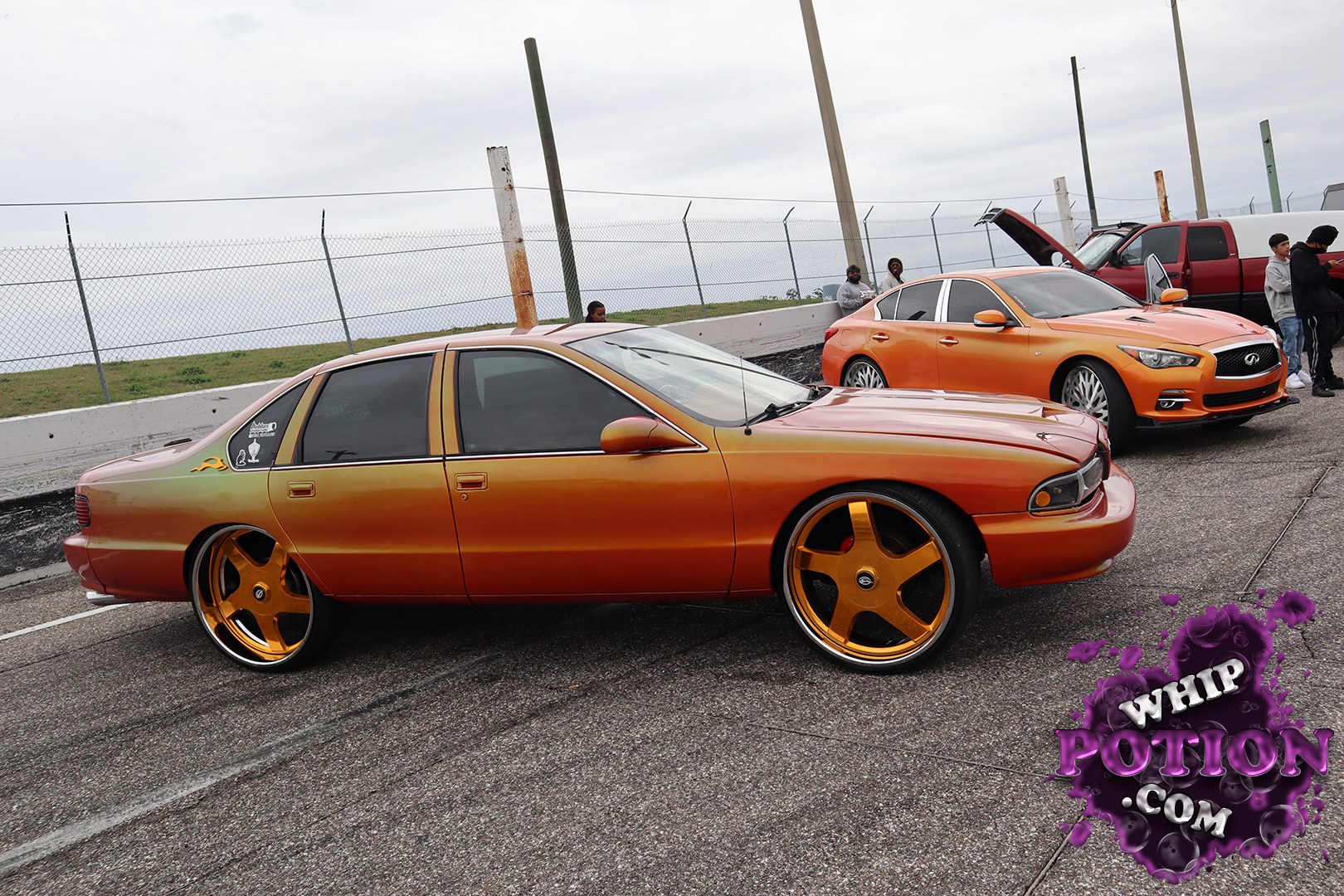 A custom four-door sedan with a deep orange-to-red fade and huge gold five-spoke wheels, sitting low and clean with a smooth tucked look. The tinted glass, shaved-down trim, and that subtle side-window sticker give it a crisp street-style vibe, while the matching orange Infiniti badge cues make the build feel thoughtfully put together.
