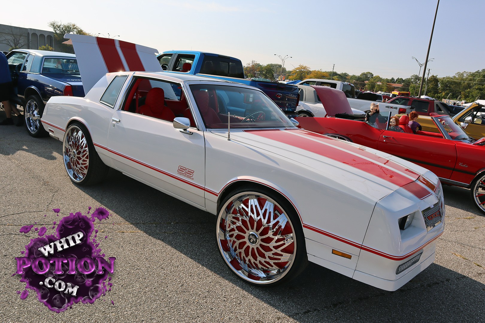 This white Chevrolet Monte Carlo SS leans hard into custom street style with its red twin stripes, matching red interior, and giant chrome-and-red spindle wheels. The shaved-looking hood and sharp two-door coupe lines give it a clean, aggressive presence, while the raised rear deck and bright trim keep the classic GM muscle-car flavor intact.