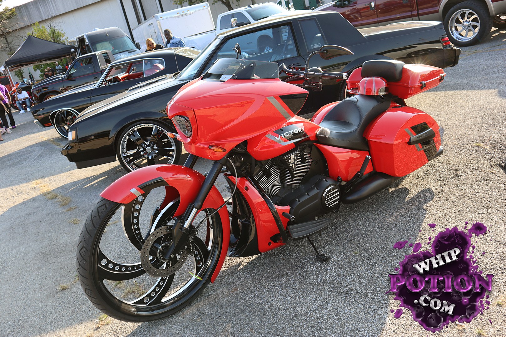 A radical custom Victory touring trike sits up front in candy red with black accents, huge billet wheels, and a long, slammed stance that gives it real show-bike presence. The matching hard bags, stretched bodywork, and exposed V-twin details make it feel more like rolling artwork than a factory cruiser.