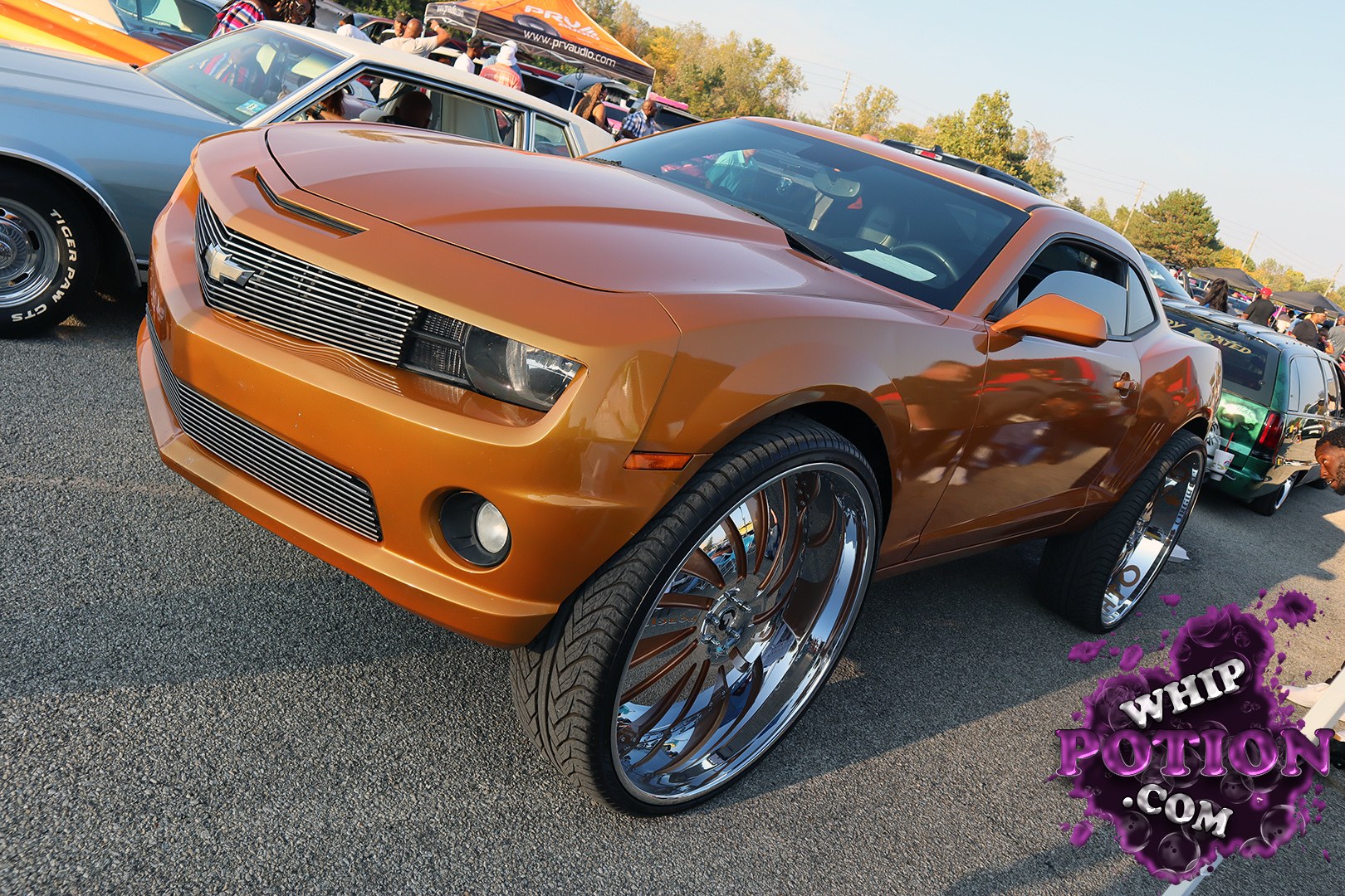This Camaro has a sharp copper-orange finish and a clean custom look, with a slim grille, smooth front fascia, and massive chrome wheels tucked under the fenders. The tall rear setup gives it that classic big-wheel show stance, while the dark tint and shaved, streamlined bodywork keep the whole build looking tight and purposeful.