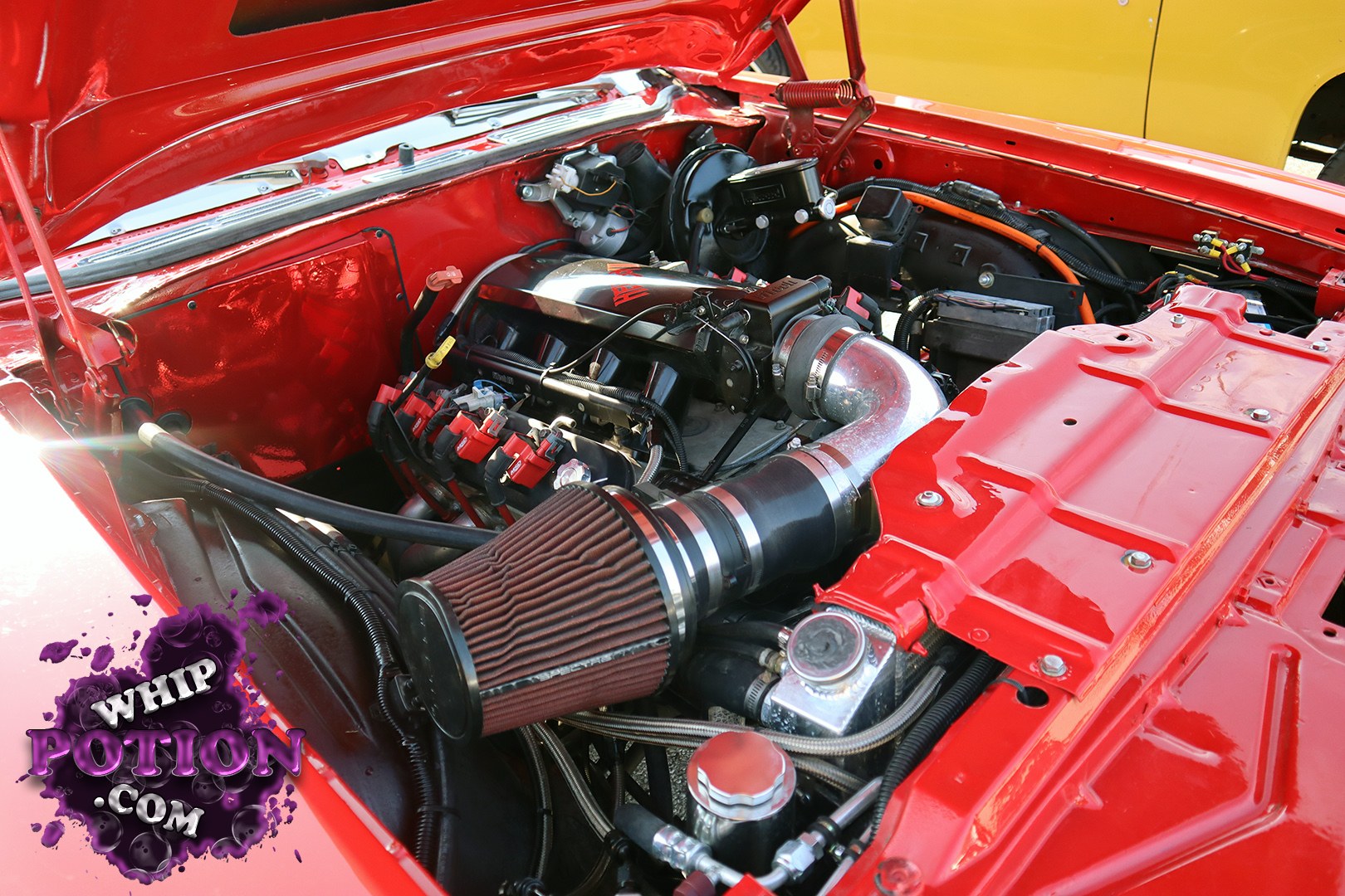 A bright red engine bay gets the full custom treatment, with a polished intake pipe, cone filter, and tidy routing that give it a clean street-machine vibe. The painted bay, upgraded ignition hardware, and neatly tucked accessories suggest a serious build that’s been finished with real attention to detail.