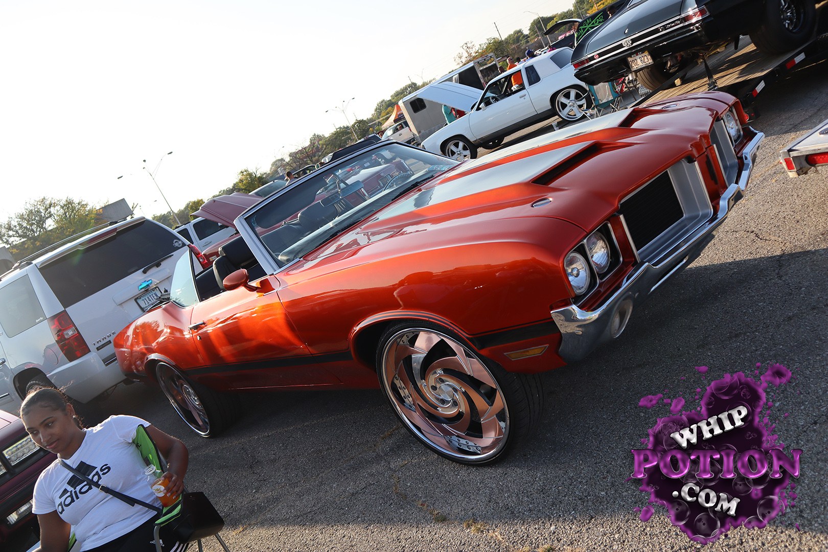 This orange Oldsmobile Cutlass convertible wears a clean custom look with deep metallic paint, a black top, and oversized chrome swirl wheels tucked under a low, ready-to-roll stance. The split grille and stacked headlights give it classic muscle-car attitude, while the smooth bodywork keeps the whole build looking sharp and deliberate.