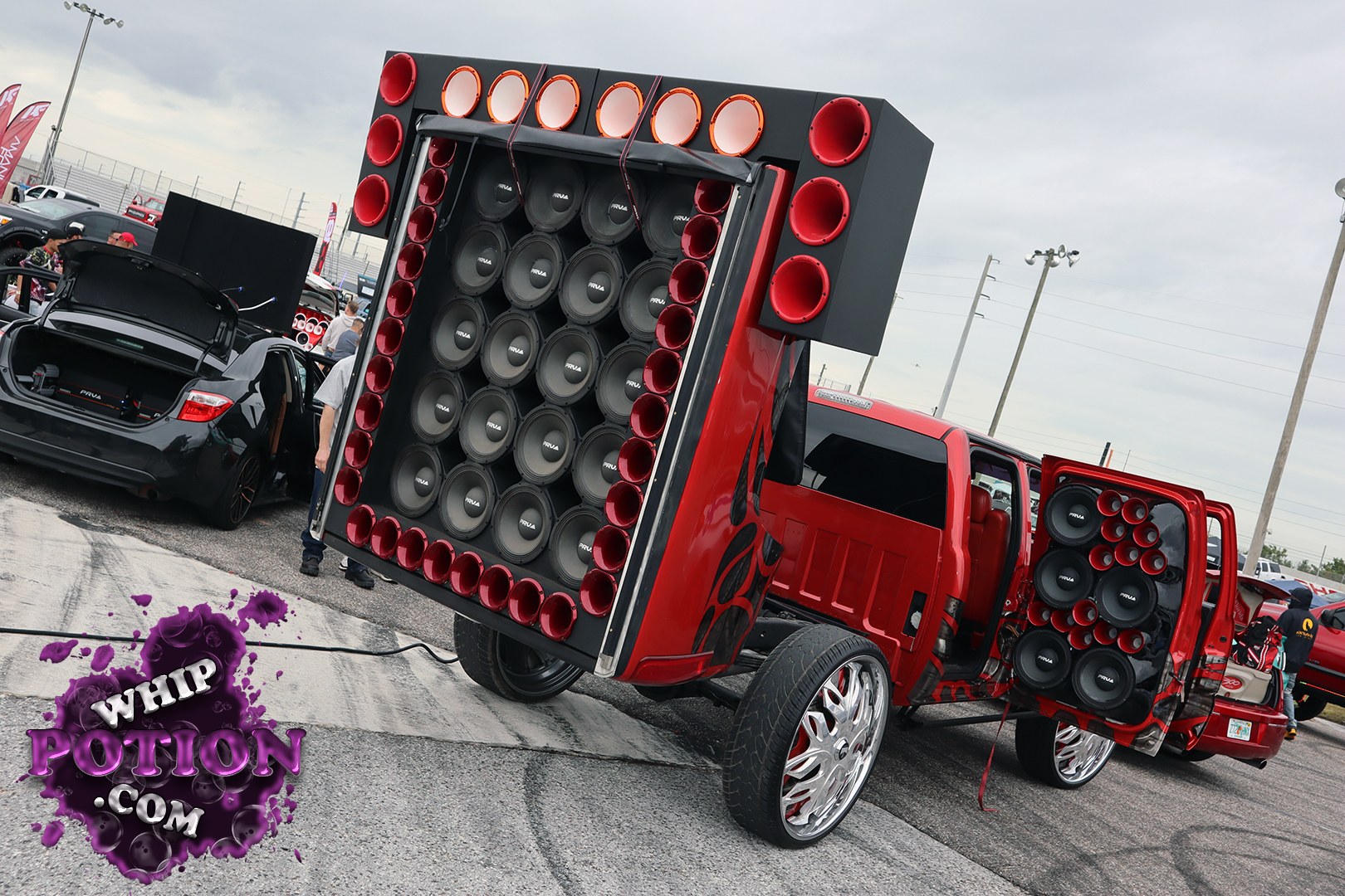 A slammed red pickup sits on oversized chrome wheels with a wild full-cab-and-bed audio wall packed with subwoofers and red horn ports. The custom build leans hard into show-truck culture, with the doors and rear hatch opened up to reveal even more speakers and a heavily modified interior trimmed to match the aggressive red-and-black theme.