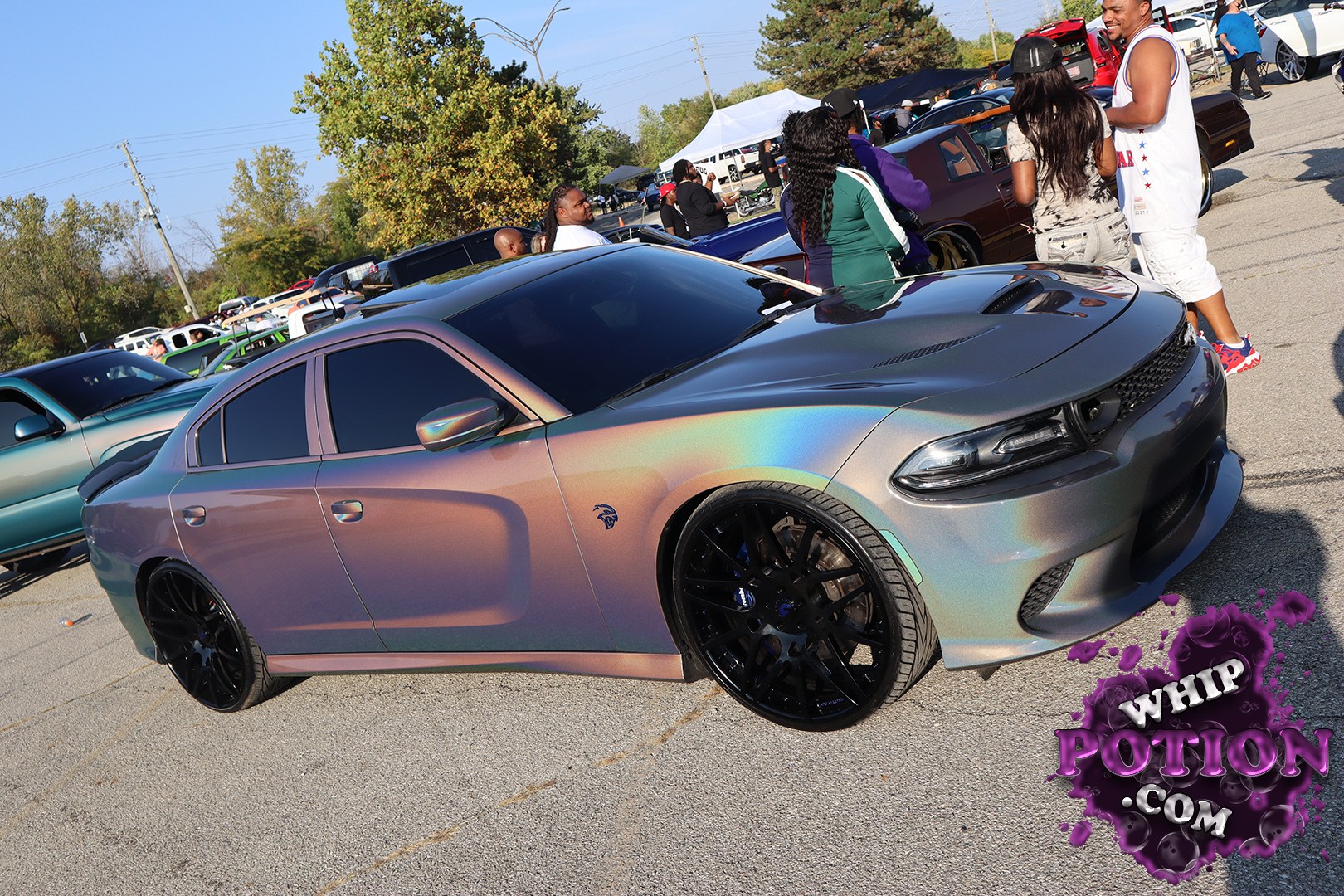 This Dodge Charger has a wild chameleon-style wrap that shifts from bronze to purple to green, paired with a gloss-black roof, tinted glass, and oversized black multi-spoke wheels. The car sits low and clean, with a subtle Demon badge on the fender and just enough stance to give it that tough street-built look without going over the top.
