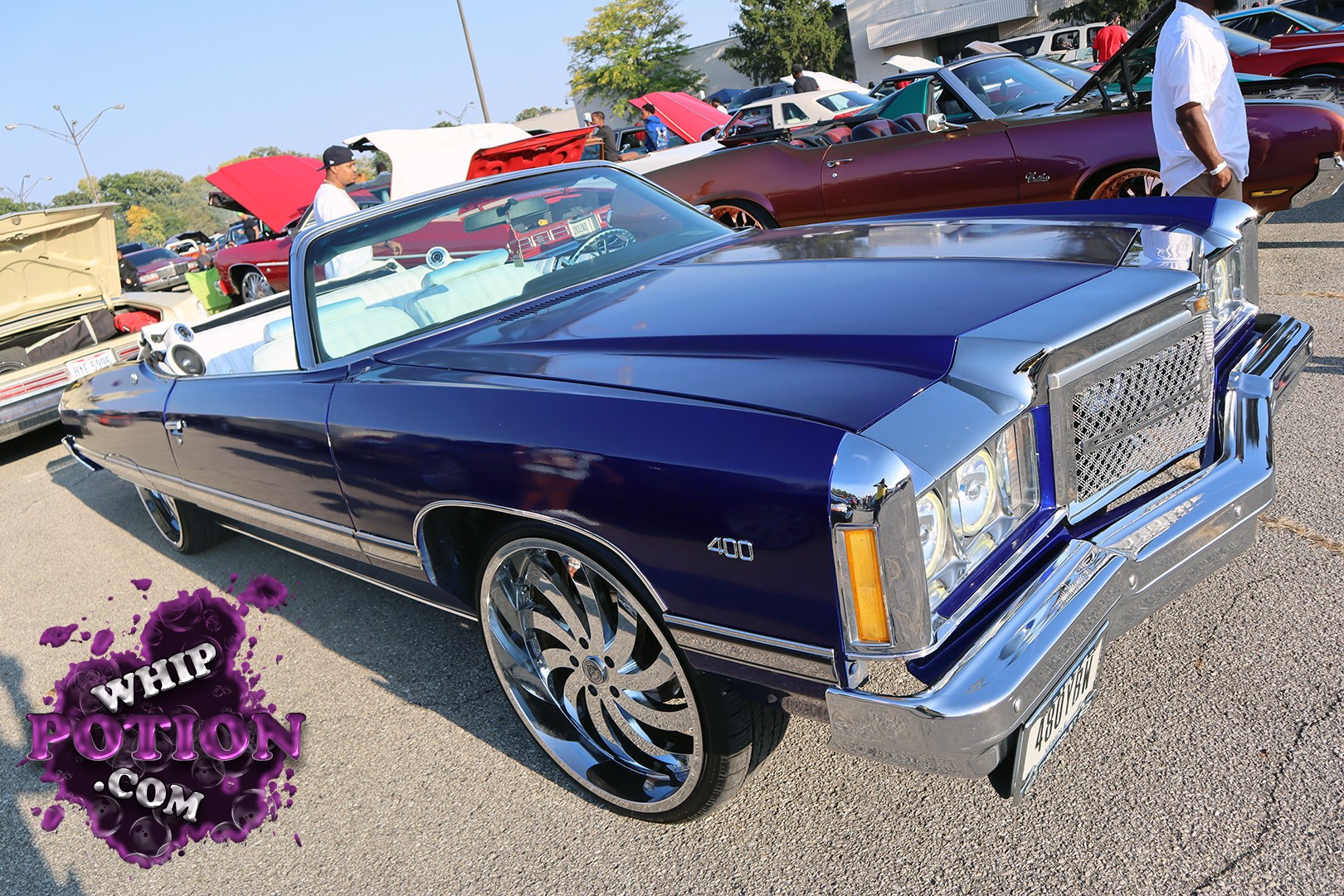 A deep metallic blue Oldsmobile Cutlass 400 convertible sits low on giant chrome spinner wheels, mixing old-school luxury with full custom swagger. The white interior pops against the paint, while the squared-off nose, bright chrome trim, and clean stance give it that well-sorted boulevard cruiser look that always gets a second glance.