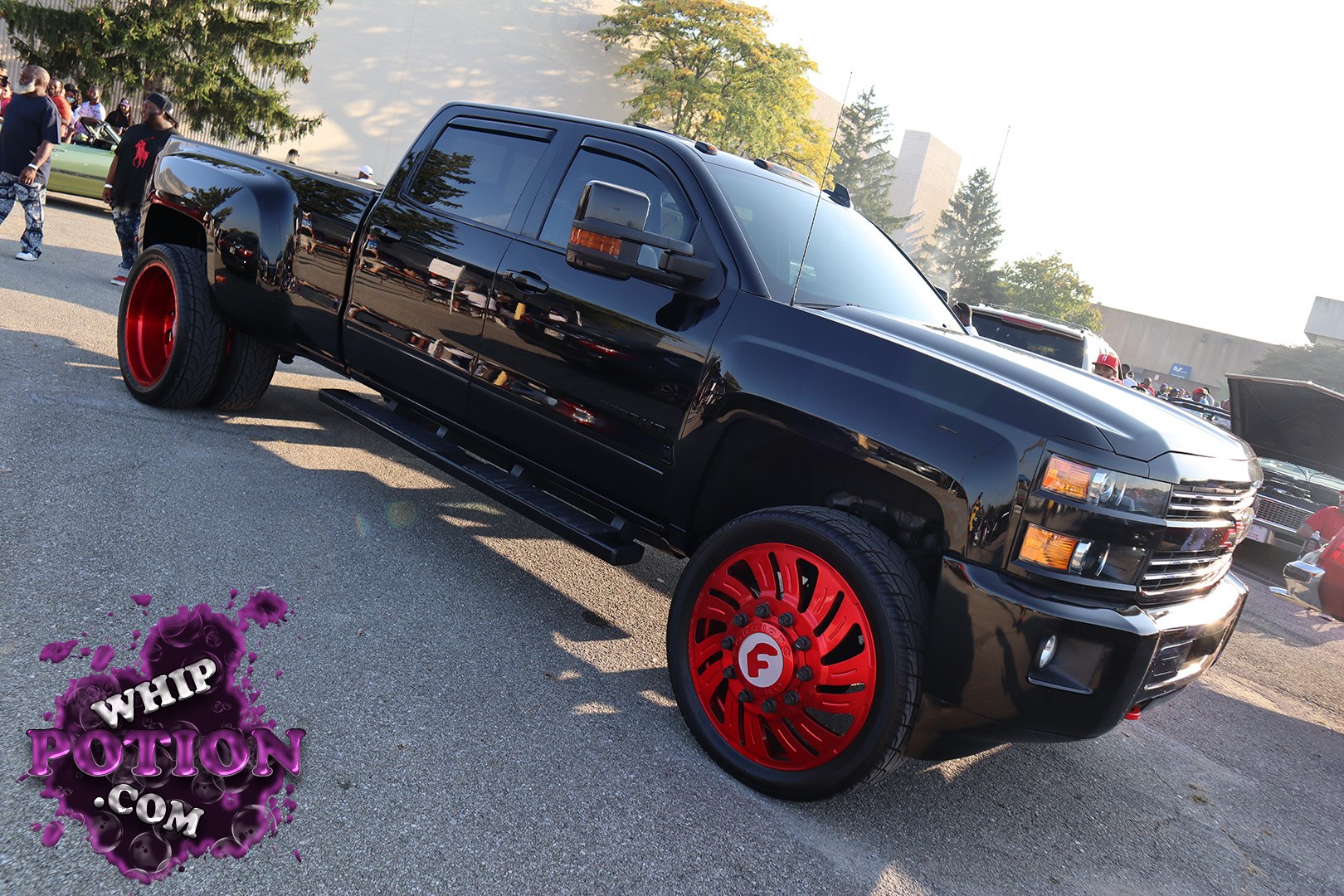 A black Chevrolet Silverado dually gets a bold custom treatment with deep red oversized wheels, a glossy murdered-out finish, and a lowered stance that gives it a serious street presence. The tinted glass, smoked trim, and clean body lines keep the look sharp, while the wide rear fenders and aggressive wheel fitment make it clear this truck was built to stand out.