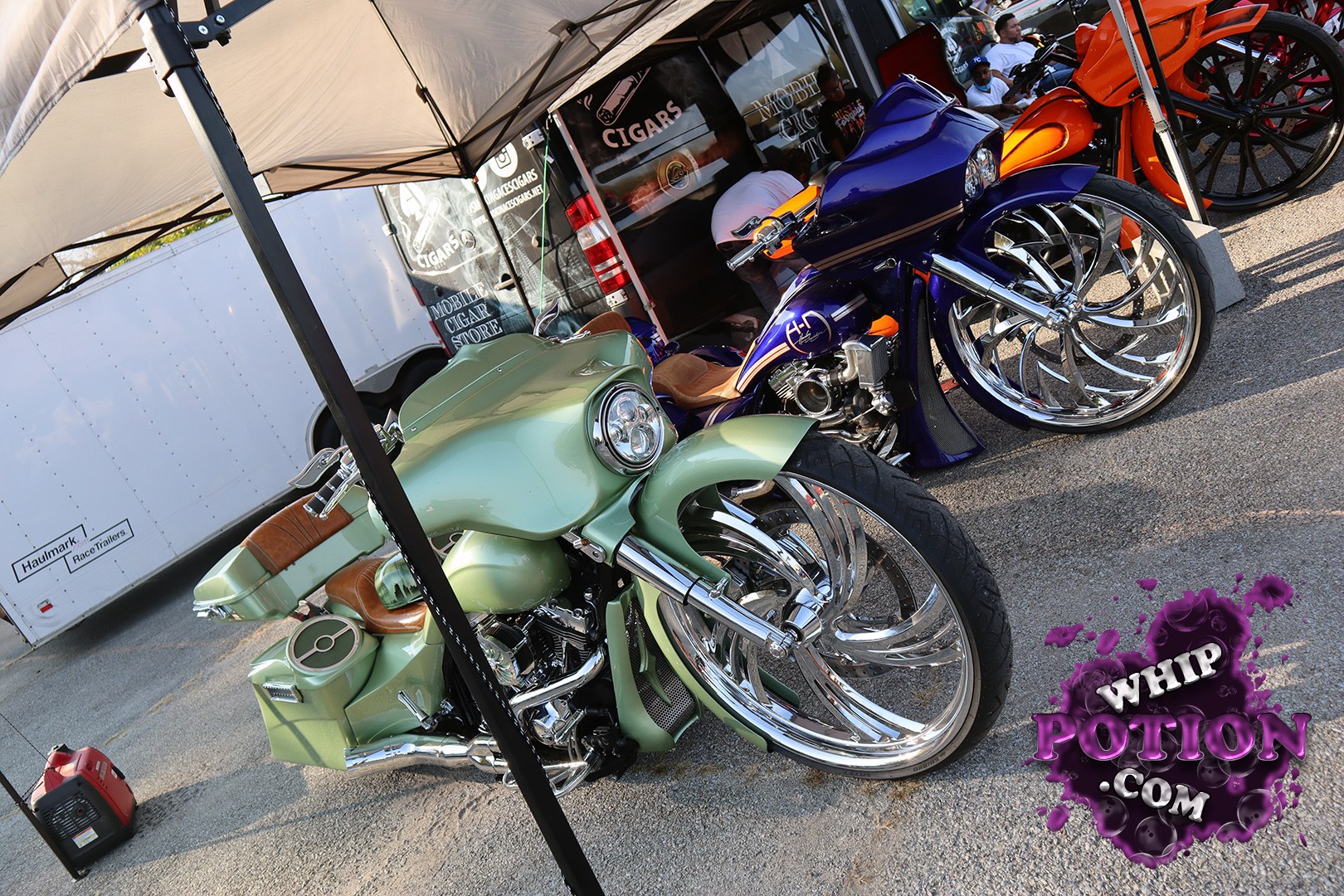 A pair of custom baggers sits under the canopy, with a mint green bike up front and a deep purple one tucked beside it, both built low with outrageous oversized chrome wheels and stretched bodywork. Tan seats, smoothed paint, and clean custom fairings give them that full-show treatment, while the orange bike in back adds another flash of color to the lineup.