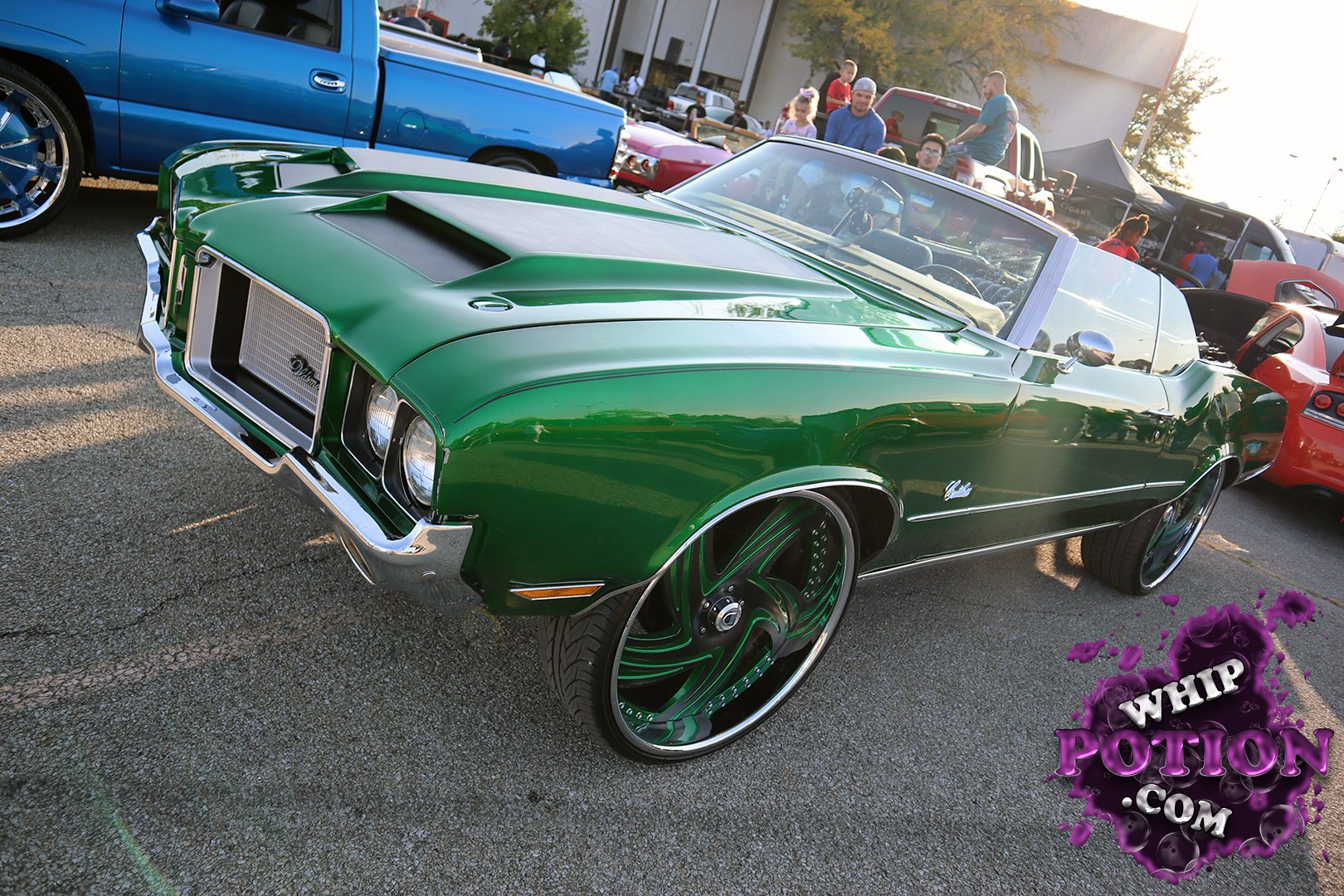 This classic Oldsmobile Cutlass convertible sits low and mean in deep metallic green, with a smoothed custom nose, shaved trim, and a blacked-out hood insert that gives it real attitude. The huge color-matched billets fill the fenders perfectly, while the clean white top and tan interior keep the whole build classy instead of overdone.