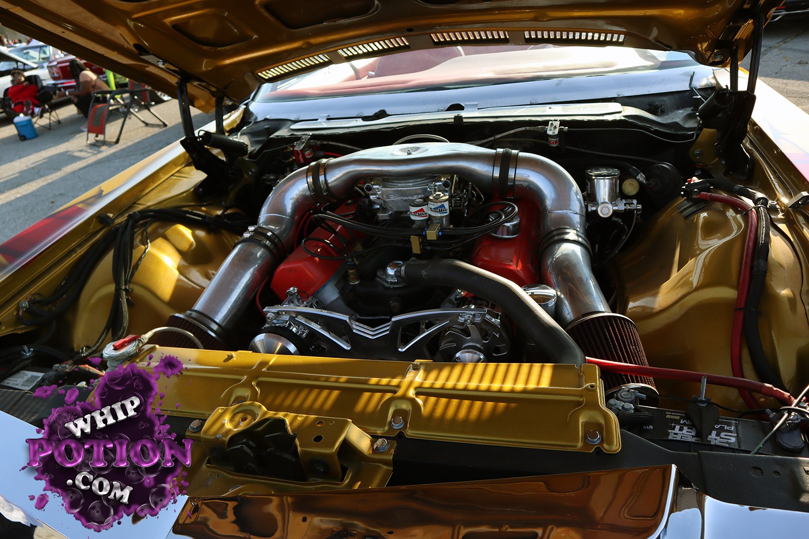 This gold-engine bay is all about attitude, with a polished intake tube sweeping over a bright red V8 and custom body-color bay work that really pops. Chrome accessory hardware, open-element filters, and the tucked plumbing give it a clean, show-ready finish that looks as serious as it is flashy.