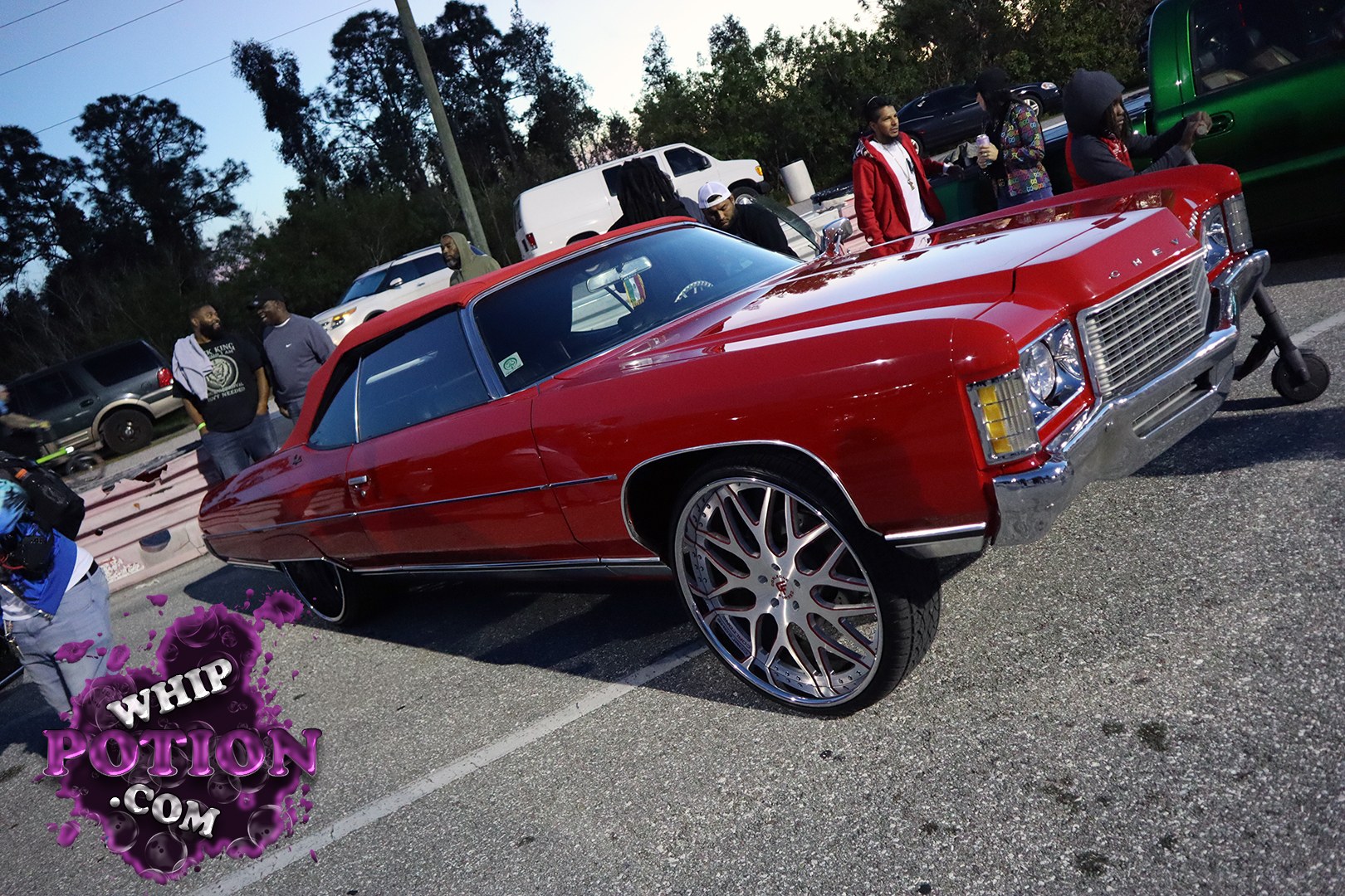 A classic Chevrolet Impala convertible sits low and clean in deep red with a matching top, long chrome trim, and a big billet wheel setup that fills the arches just right. The front end keeps that ’70s Chevy presence with stacked lamps, a wide grille, and plenty of polished metal doing the talking.