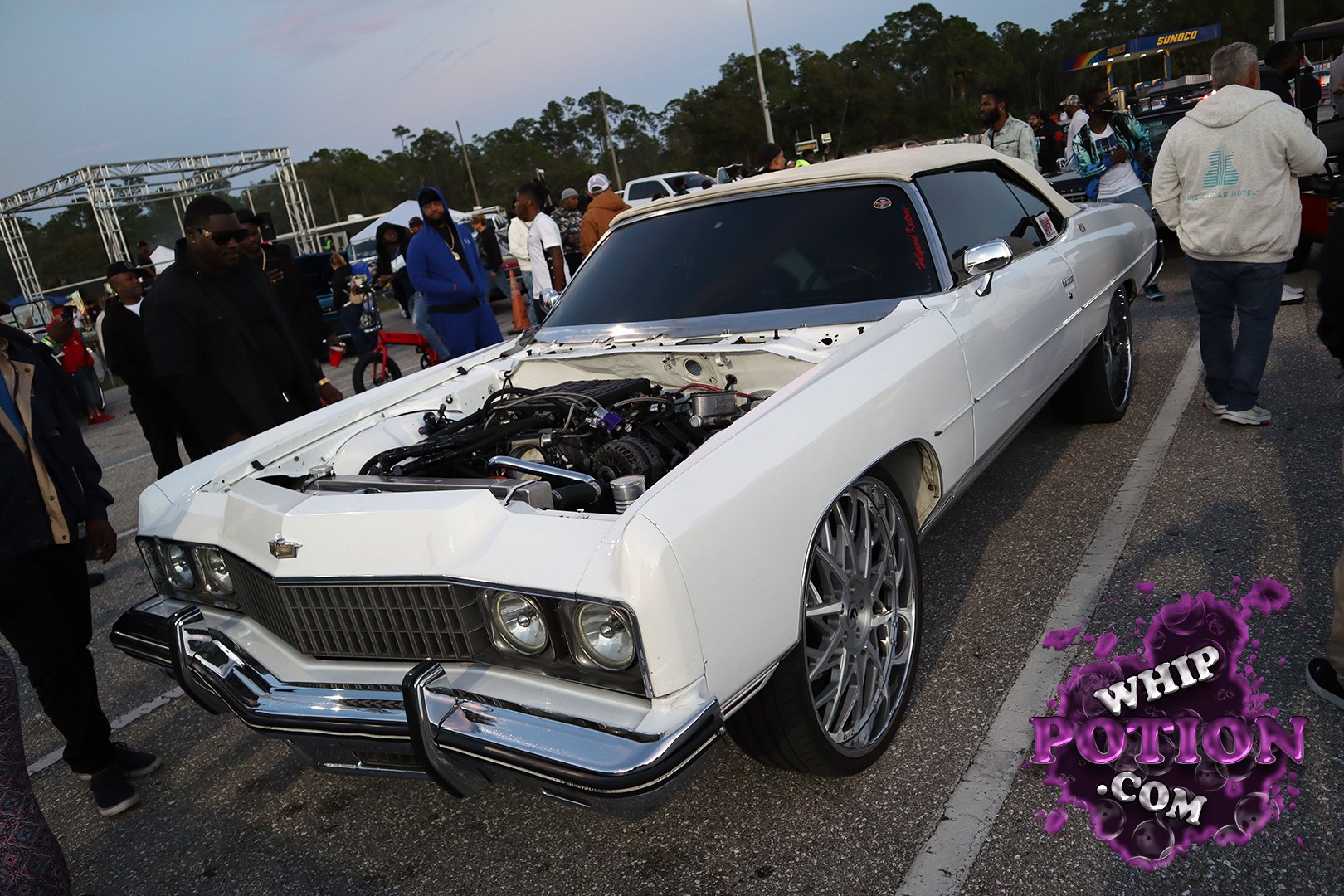 This old-school Cadillac hardtop has real presence with its shaved, clean white body, blacked-out glass, and massive staggered chrome-and-mesh wheels tucked under the fenders. The hood’s off to show a tidy engine bay, while the long, low profile and bright bumper work give it that classic cruiser attitude with a modern custom touch.