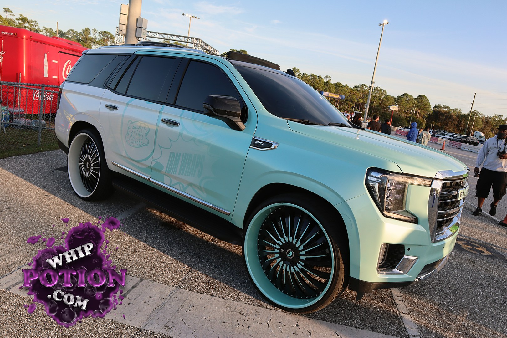 This GMC Yukon goes full custom with a mint-and-white two-tone wrap, matching painted wheels, and deep-tinted glass that gives the big SUV a clean, upscale look. The oversized black-and-mint rollers and lowered stance bring real show-truck attitude, while the “IBM Wraps” and cancer awareness graphics add a personal touch.