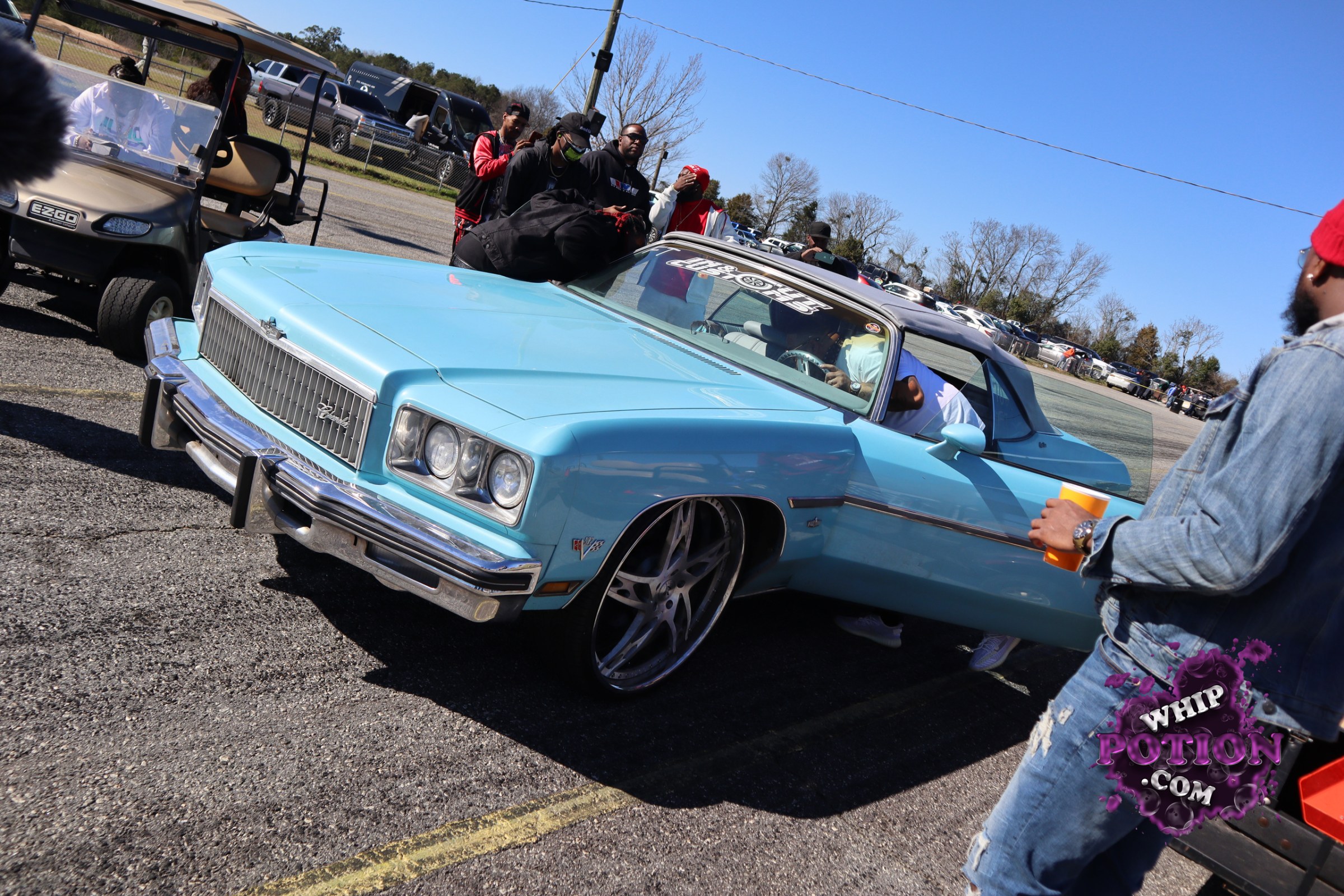 A sky-blue Chevrolet convertible cruises in with serious presence, its chrome grille, quad headlamps, and long body lines giving it that classic mid-’70s GM swagger. Oversized polished wheels tucked tight under the fenders and the low stance give it a clean custom look, while the black top and matching trim keep the build nicely balanced.