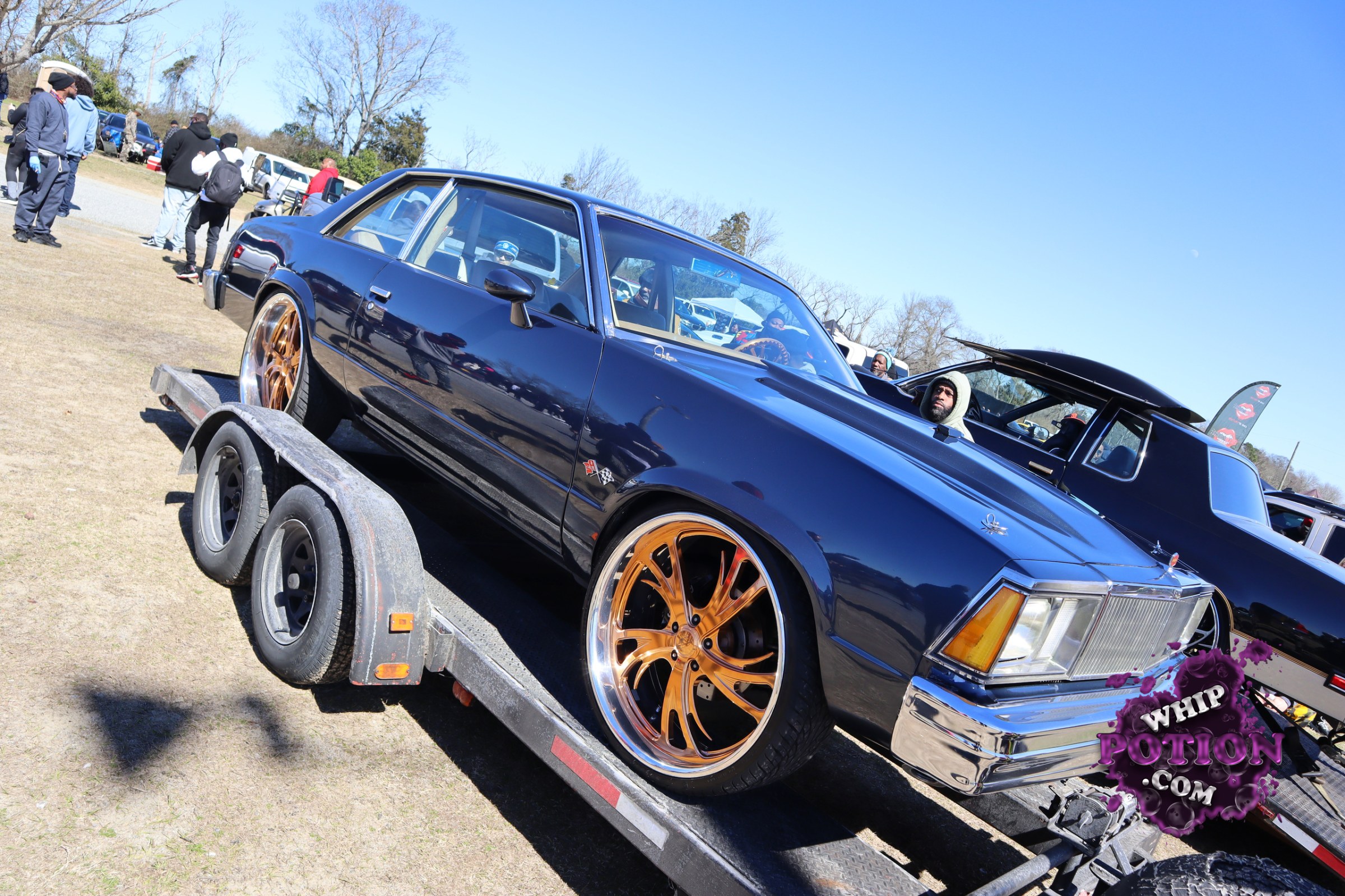 A dark blue classic coupe sits low on oversized bronze-and-chrome wheels, with a clean chrome grille, rectangular headlights, and a long, squared-off body that gives it serious old-school presence. The white interior peeks through the glass, and the polished trim plus big rear rubber make this one look ready for both cruising and showing off.