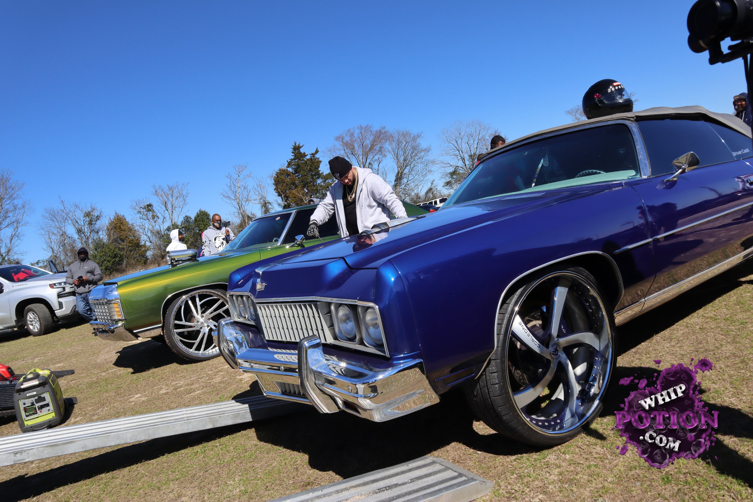 A pair of big-body classics sit nose-out with full custom attitude, led by a deep purple coupe with a razor-sharp front clip, heavy chrome, and oversized billet wheels tucked under a low, clean stance. Beside it, the metallic green-and-gold car leans into the same street-luxury vibe with giant wheels, polished trim, and a long, flowing profile.