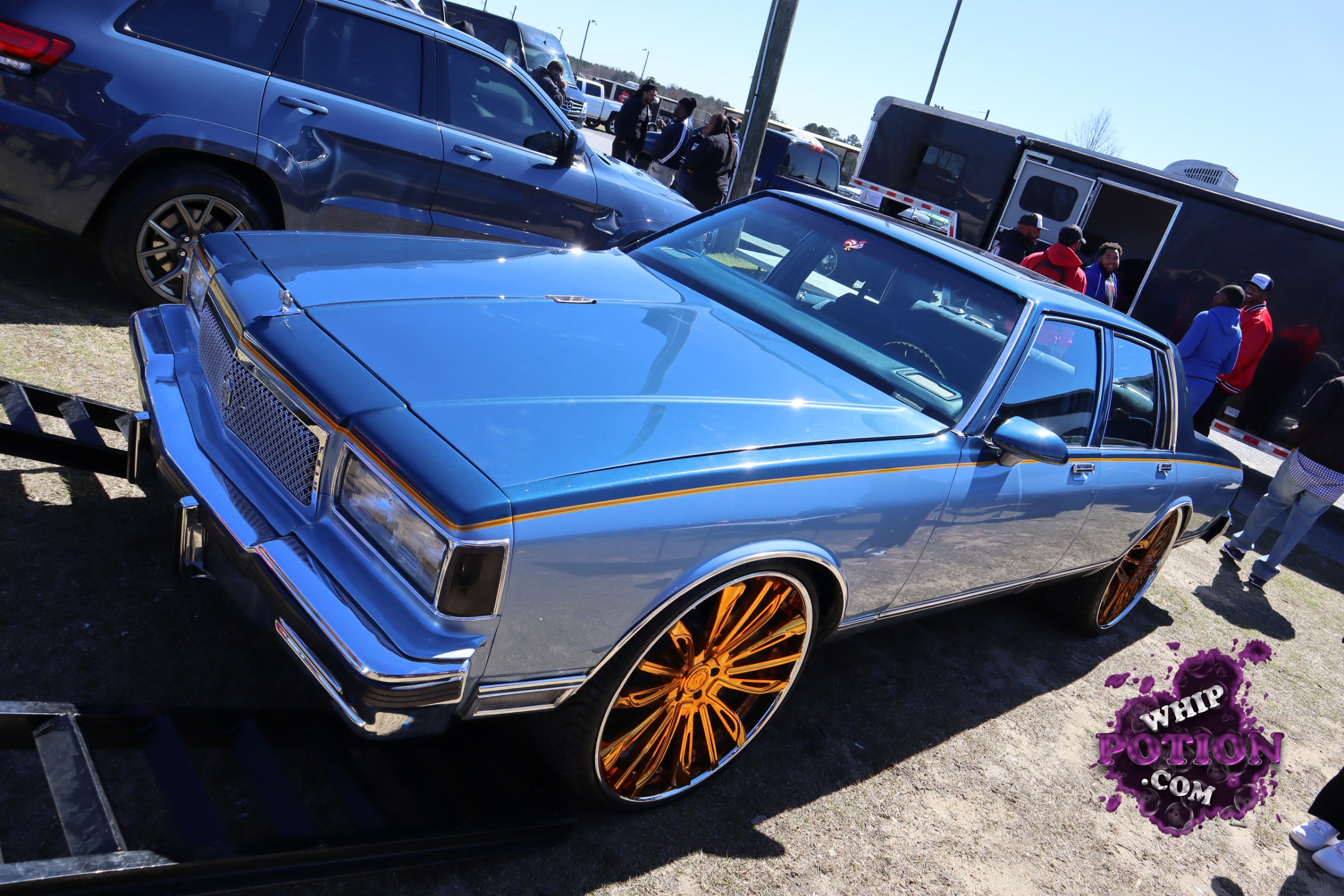 A blue boxy Buick sedan sits low on oversized gold-dipped rims, with a clean two-tone-style trim strip, chrome grille, and long straight body lines giving it that classic old-school presence. The stance is aggressive without going overboard, and the glossy paint really pops against all that polished wheel hardware.