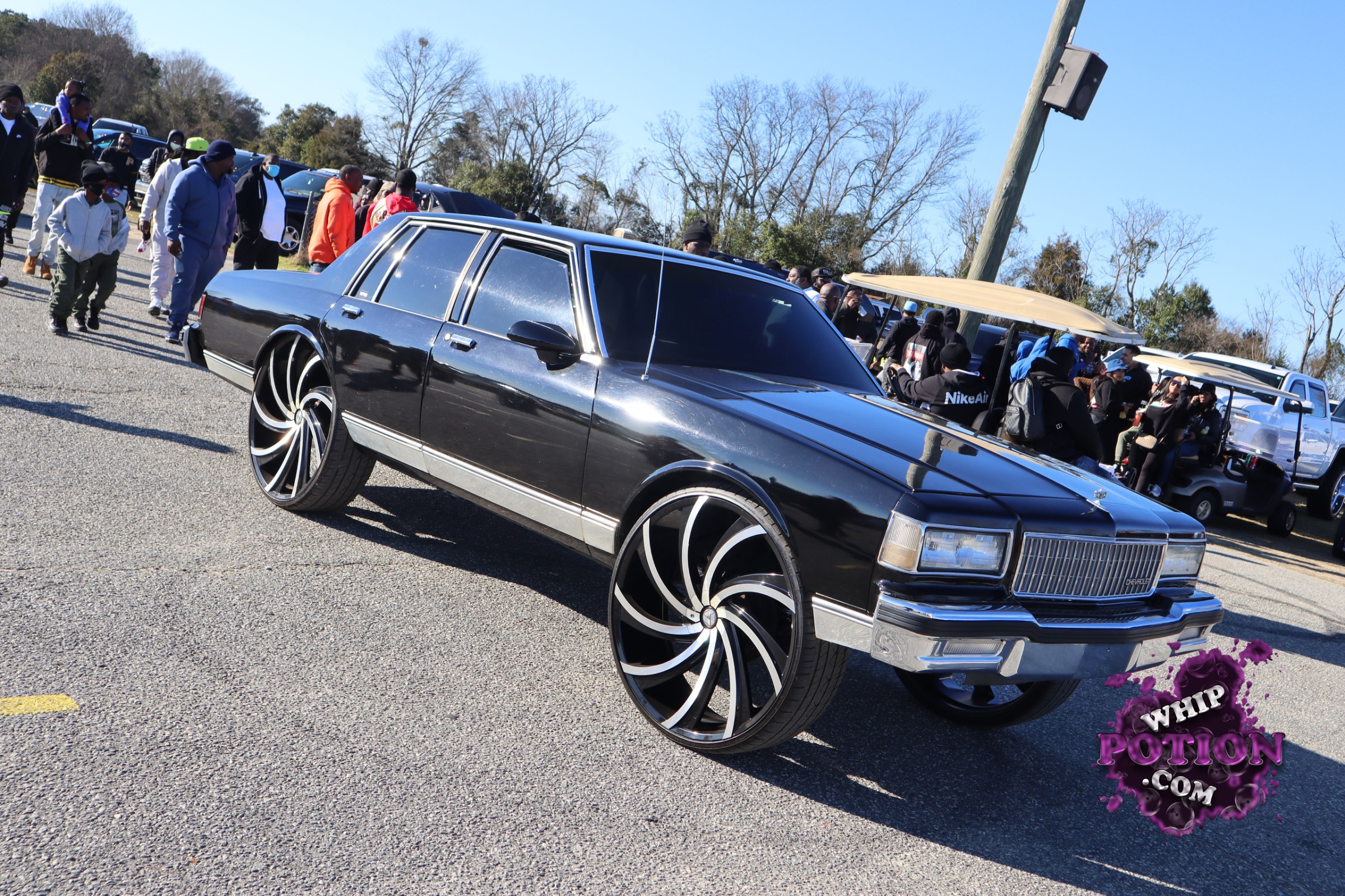 A boxy old-school GM sedan sits nose-down on massive black-and-chrome wheels, with a clean black paint job, heavy tint, and bright chrome trim keeping the classic lines crisp. The grille badge points to a Chevrolet Caprice, and the oversized rollers give it that hard-hitting custom street presence without losing its original full-size attitude.