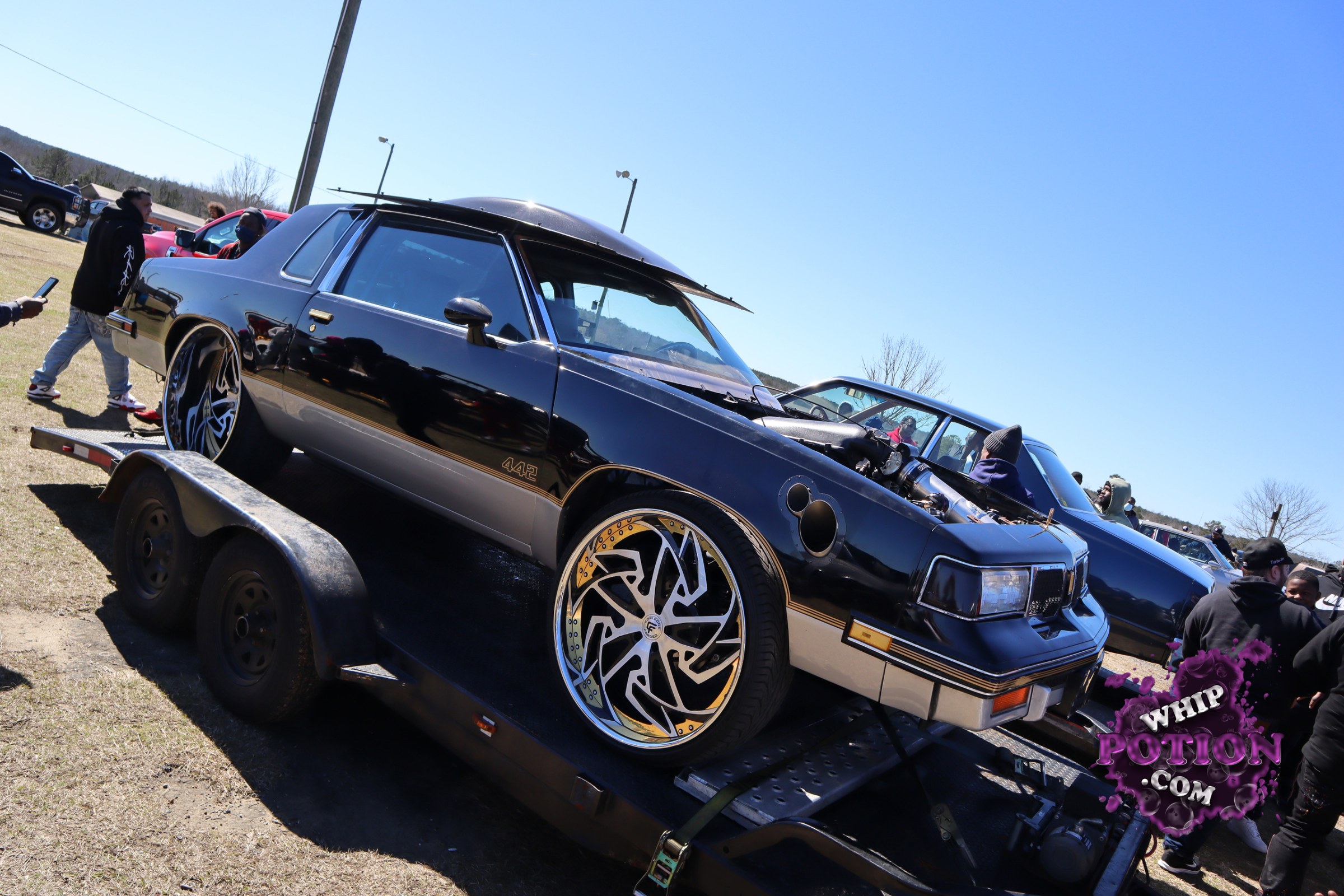 A sharply built Buick Regal 442 sits low on a trailer, wearing glossy black-and-silver two-tone paint, massive custom multi-piece wheels, and a gold-trimmed side stripe. The hood’s been swapped for a raised, cutaway-style setup that shows off the engine hardware, giving this classic G-body a serious street-and-show attitude.