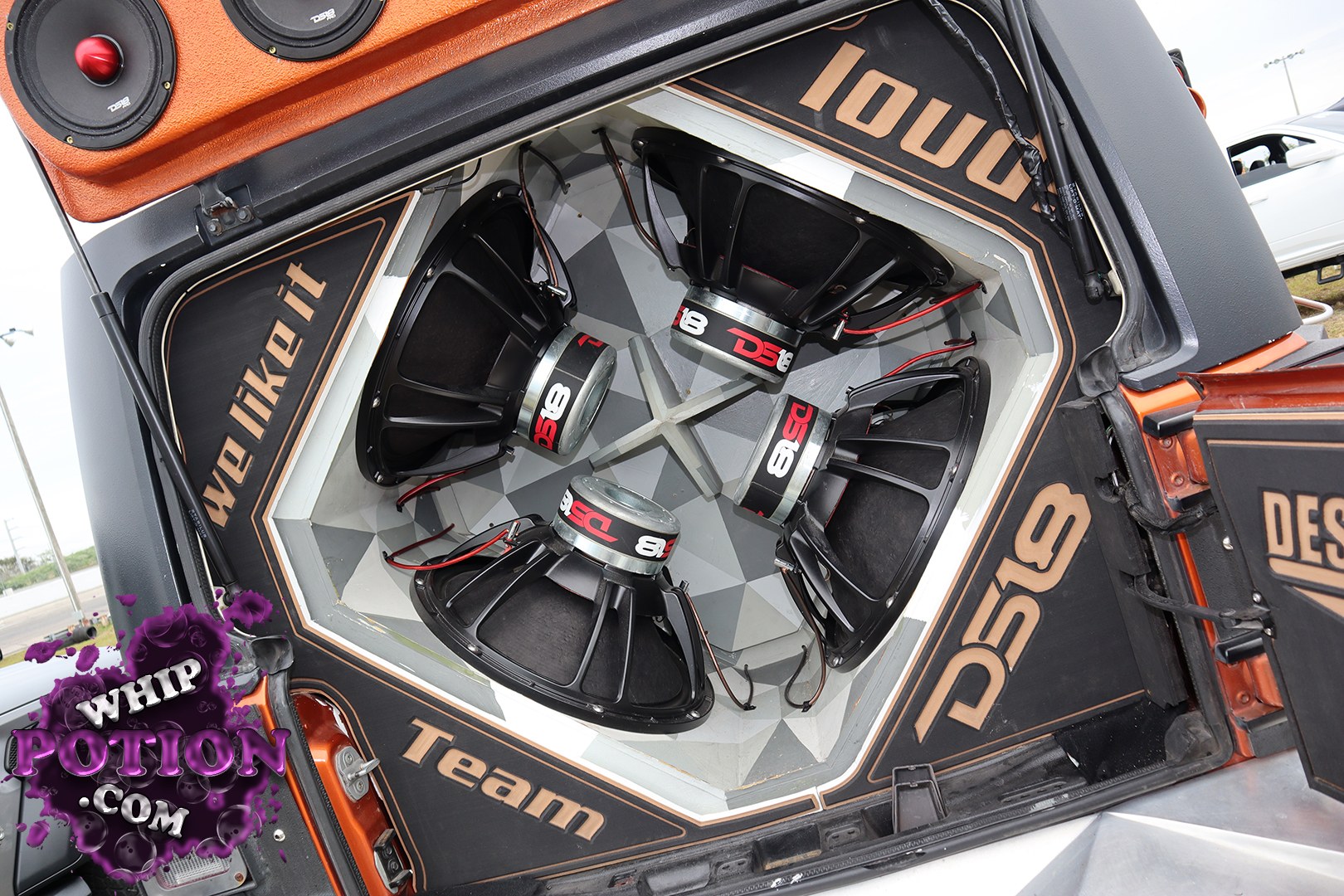 This custom audio build is all about the trunk, with four massive DS18 subwoofers sunk into a clean, faceted enclosure and framed by orange-and-black panels with bold “DS18” and “Team” lettering. The install has a serious show-truck vibe, mixing sharp fabrication, tight wiring, and a full wall of bass-ready hardware.