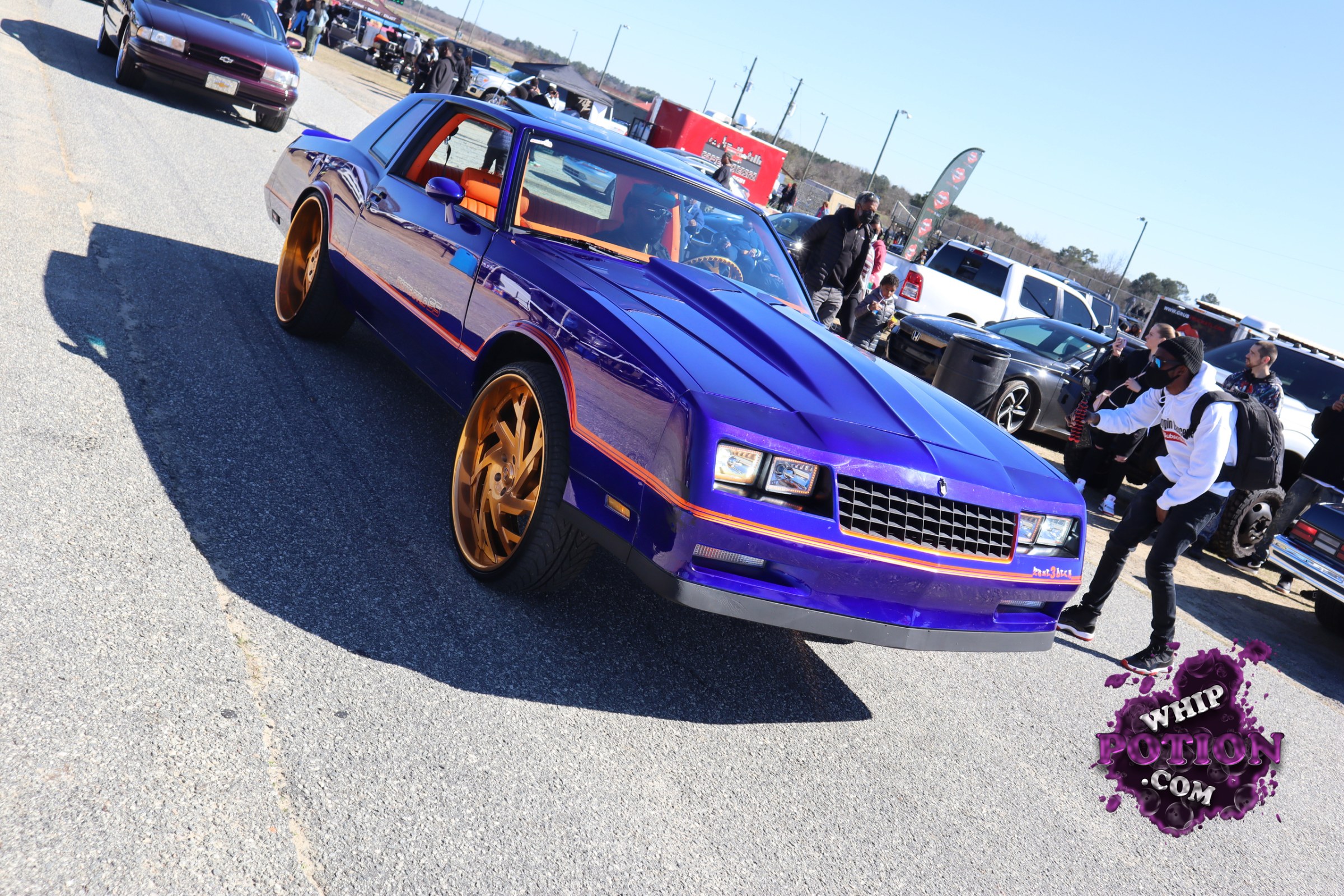A deep violet Chevrolet Monte Carlo gets the full custom treatment here, with a slammed stance, oversized gold wheels, and a bright orange interior that really pops through the glass. The clean body lines, hood bulge, and crisp striping give it that classic street-machine attitude, finished off with a look that’s equal parts show car and boulevard bruiser.