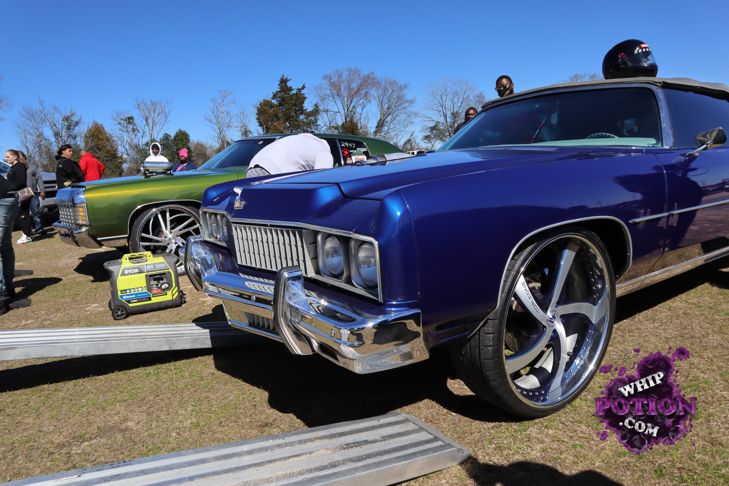 A pair of old-school American cruisers sit low and proud here, led by a deep blue convertible with a white top, huge custom five-spokes, and a heavily chromed nose. Beside it, the green hardtop wears matching oversized rollers and a clean two-tone vibe. Both cars lean into that classic big-body boulevard style with modern wheel fitment and serious presence.