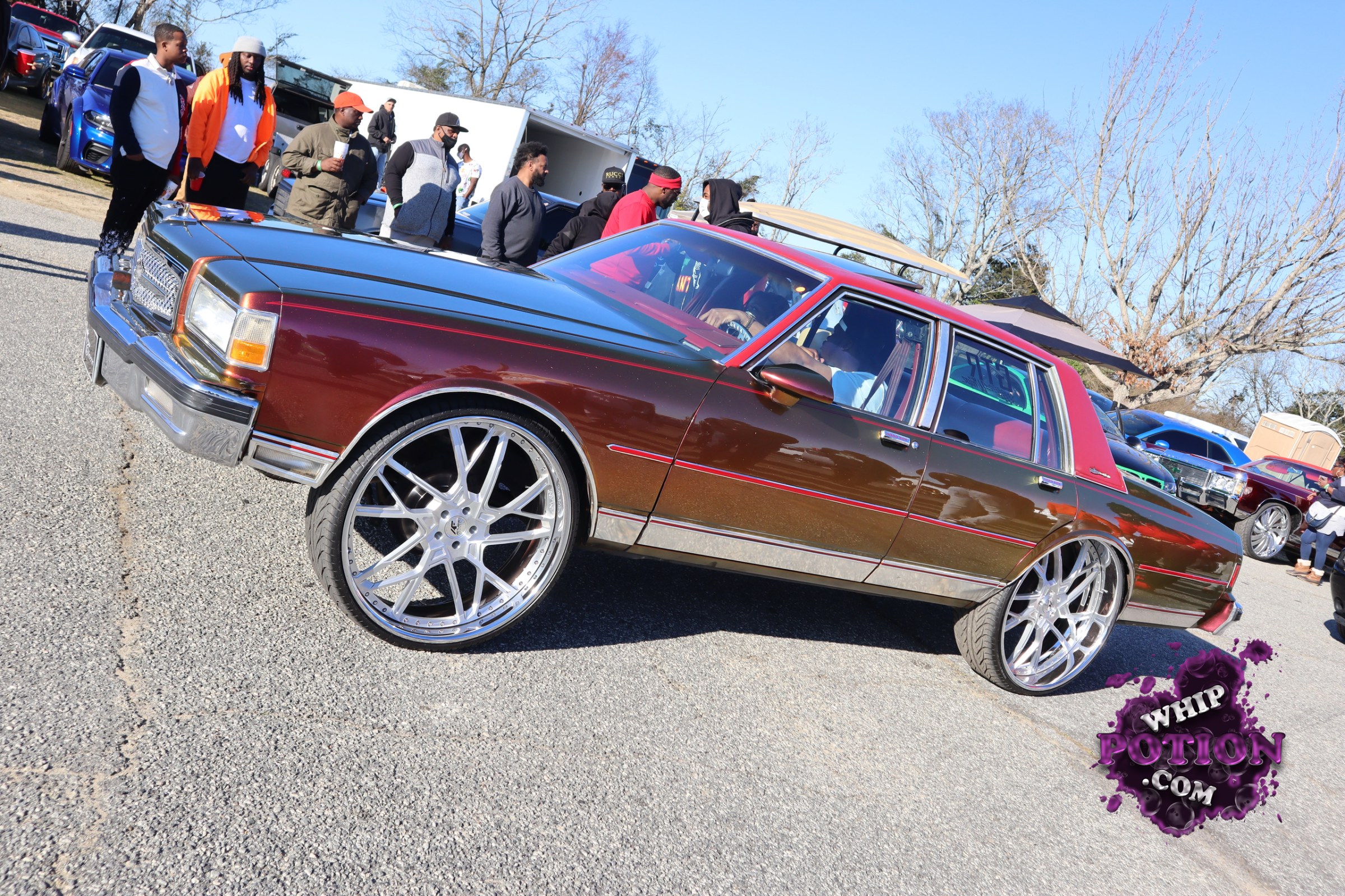 A heavily customized classic Cadillac sedan with a deep burgundy and brown two-tone finish, a black hood, and matching red roof inserts. The big billet-style wheels fill the wells nicely, giving it a clean, aggressive stance, while the chrome trim and squared-off body lines keep that old-school luxury vibe intact.