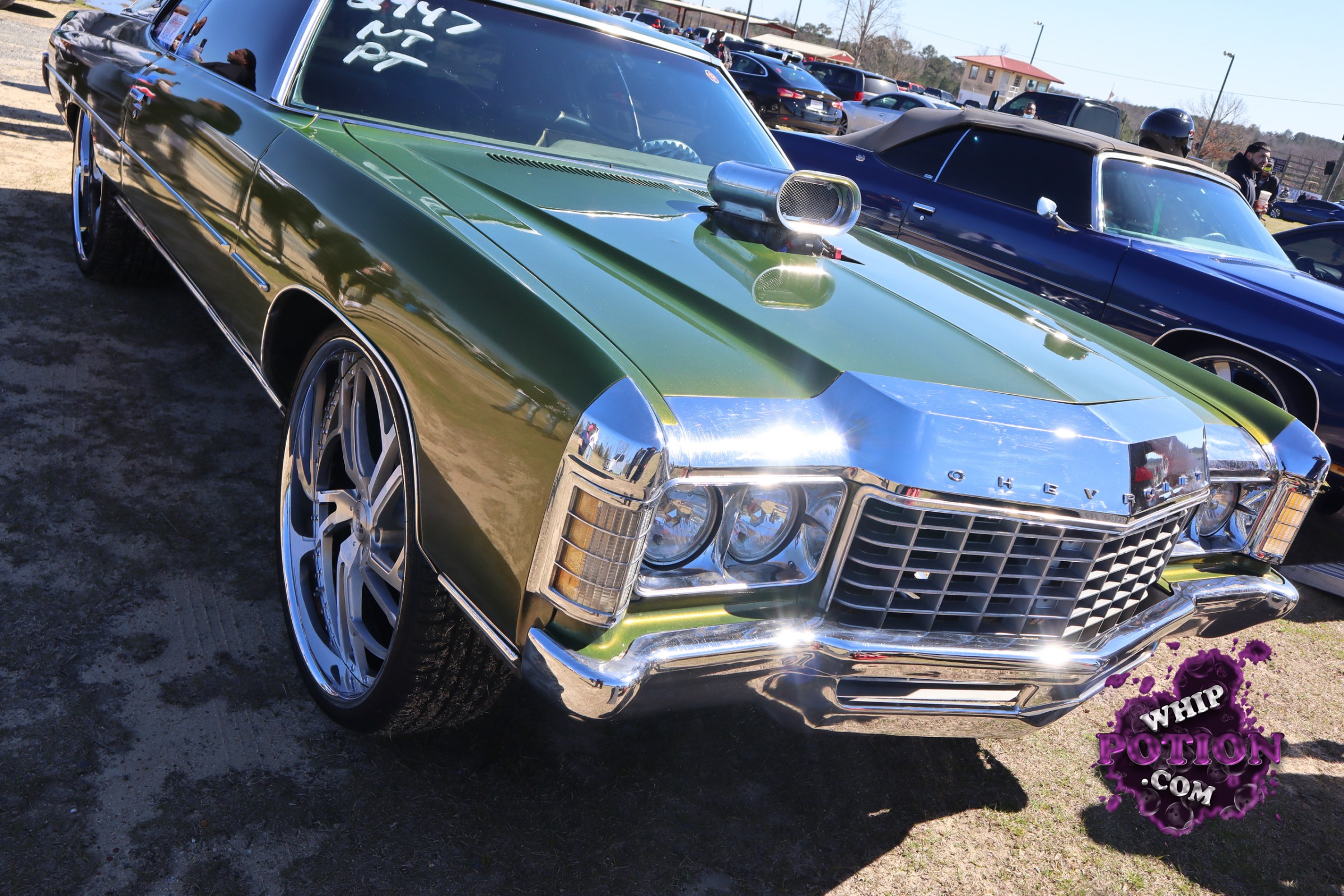 A slammed late-’60s Chevrolet coupe in metallic green gets all the right street-cruise attitude with huge chrome wheels, thin rubber, and a polished blower sticking through the hood. The twin-square front end, stacked headlights, and broad chrome grille give it that unmistakable full-size Chevy presence, while the mirror-finish trim and aggressive stance make it feel every bit the showpiece.