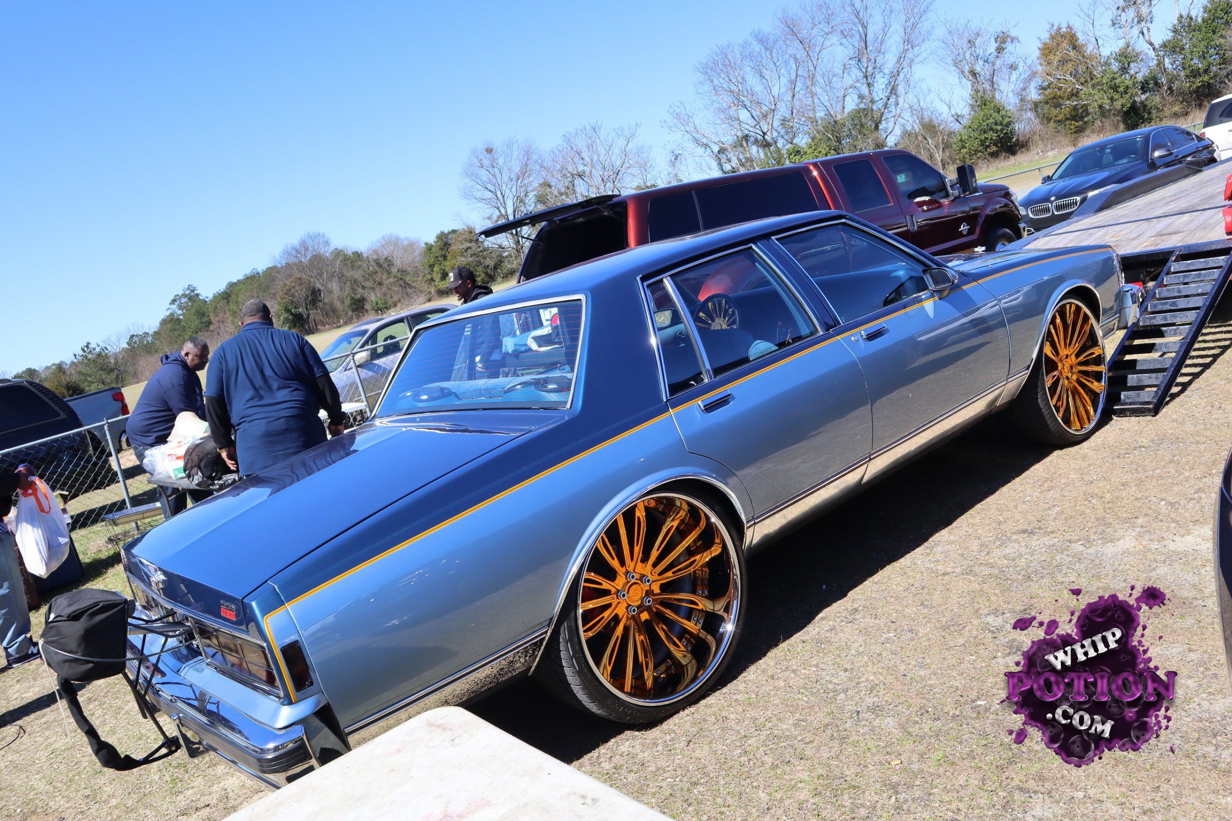 A clean, boxy full-size sedan with classic Cadillac styling sits low on huge gold multi-spoke wheels, the blue-and-gray two-tone paint sharpened by a thin gold pinstripe. The chrome bumpers and long, formal roofline give it a proper old-school boulevard look, while the stretched stance and deep-lipped rims bring plenty of custom flavor.