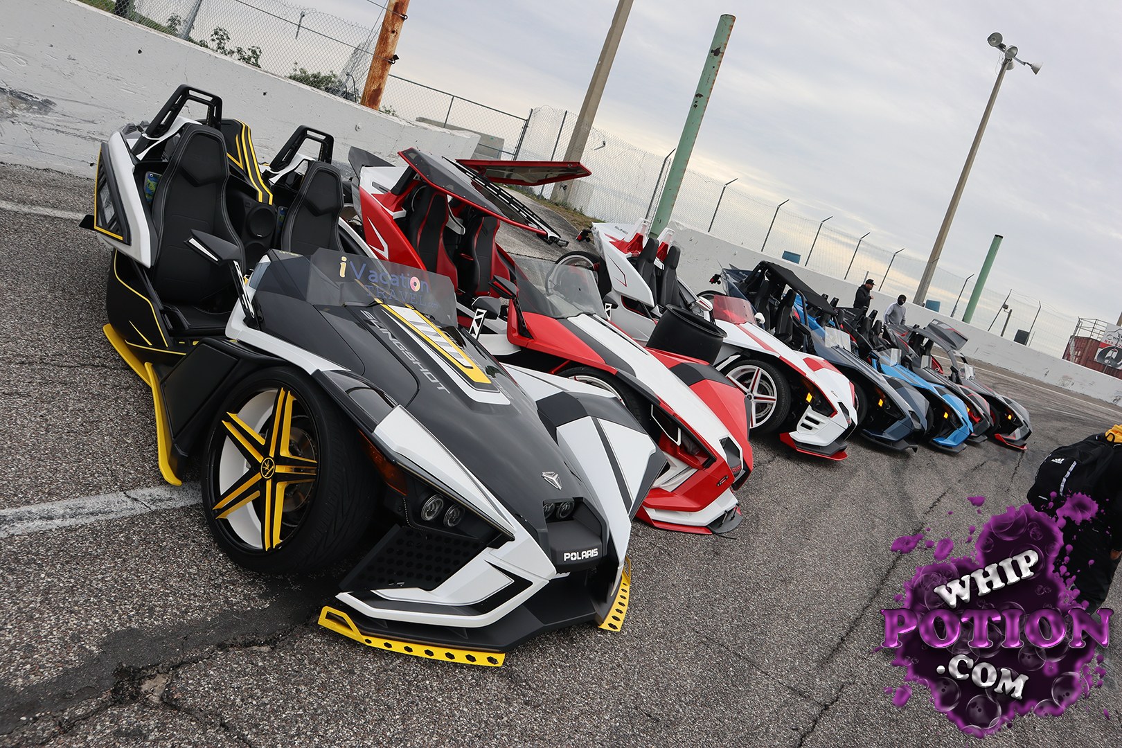 A row of low-slung, open-cockpit Slingshots sits angled across an industrial asphalt lot, each dressed in bold custom wrap schemes of black, white, red, blue, and gray. The nearest machine features yellow-accented wheels, sharp angular bodywork, and an aggressive nose close to the ground. Overcast light softens the scene, while the fenced backdrop and concrete barriers give the lineup a gritty, motorsport-showcase atmosphere.