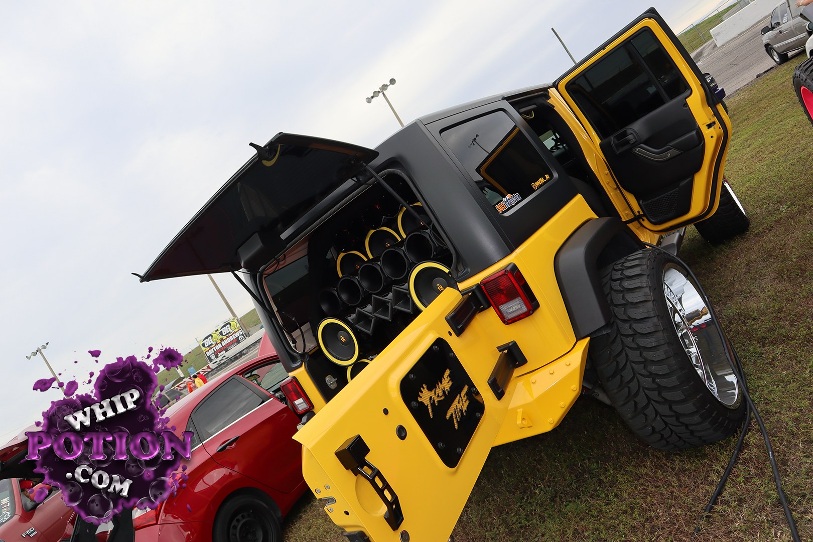 This Jeep Wrangler has been turned into a full-on show build, with bright yellow bodywork, a black hardtop, and the rear packed tight with a massive wall of subwoofers and horn-style speakers. The oversized polished wheels and chunky off-road tires give it a wild stance, and the open doors let you see the custom interior touchpoints that tie the whole setup together.