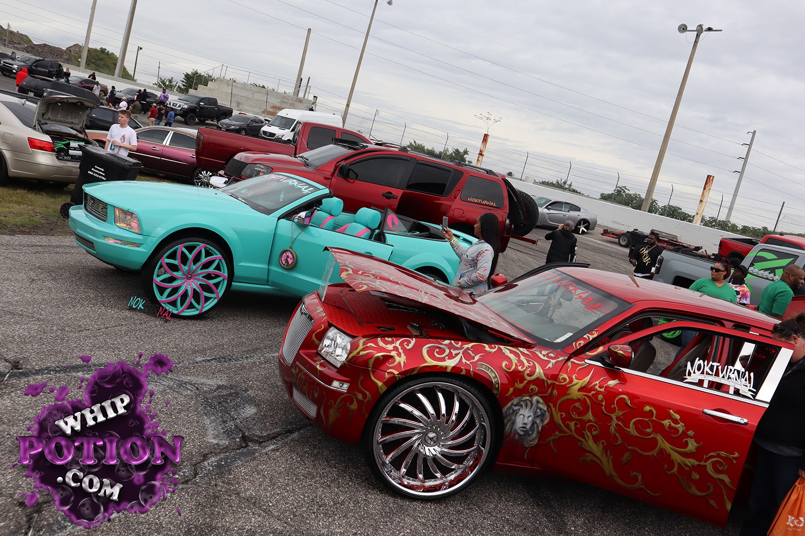 A turquoise convertible Mustang sits low on oversized turquoise-and-pink wheels, with a matching custom interior that keeps the whole build cohesive. In front, a red sedan wears wild gold pinstriping, a chrome billet-style wheel package, and a heavily customized look with the hood popped for display. Both cars bring real show-floor personality.