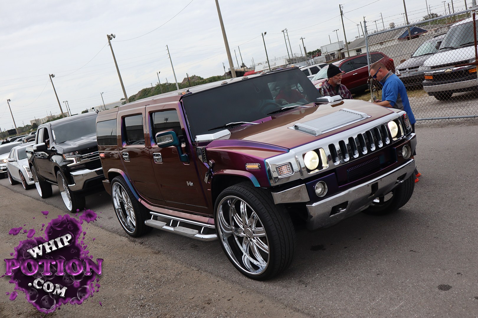 A Hummer H2 gets a full custom treatment here, wearing a deep candy-style purple-and-bronze finish with chrome-heavy trim, a vented hood insert, and huge polished multi-spoke wheels tucked under the fenders. The stance sits just right for a show build, and the mirror-like wheels and side steps give it that bold, upscale street-cruiser look.