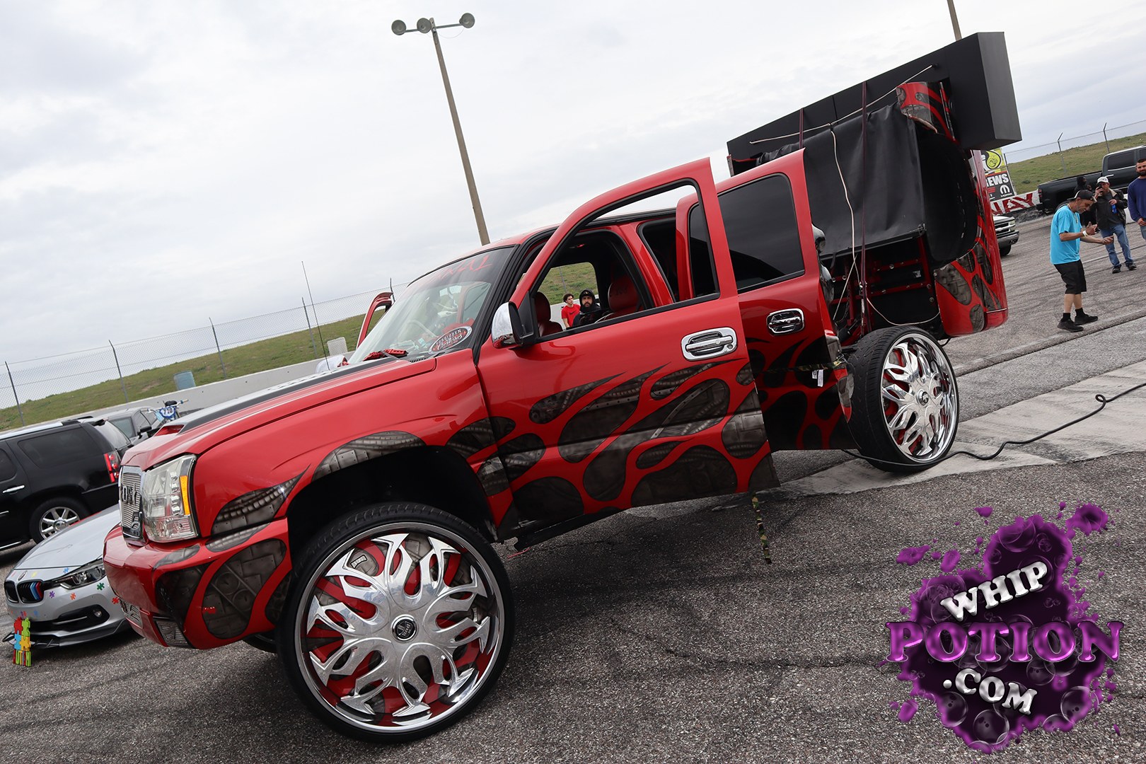 This lifted red GMC pickup is built loud in every sense, with massive chrome billet wheels, black flame-style graphics, and a wild custom bed setup packed with audio gear. The doors are open to show off the red interior, while the towering rear box and oversized stance give it that unmistakable show-truck attitude.