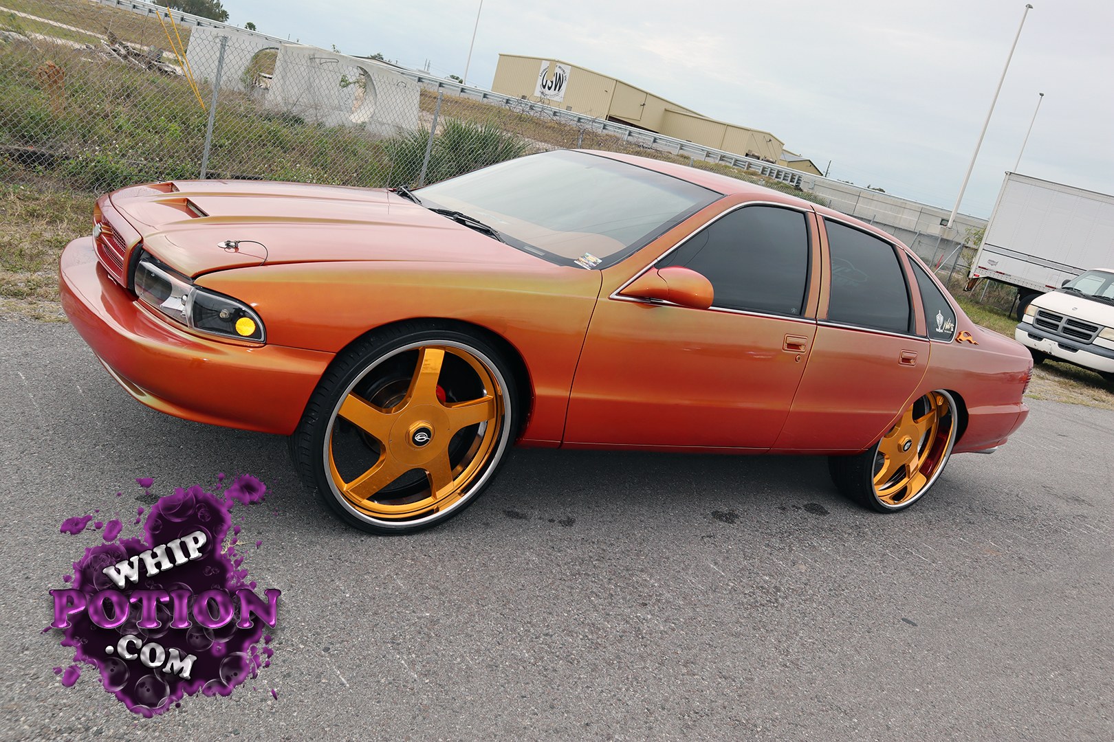 A slammed Buick four-door sedan gets a full candy fade from copper to deep red, sitting over massive gold five-spokes with the fenders tucked tight. The shaved, monochrome look keeps it clean, while the tinted glass and smooth bumper treatment give it that properly built street-cruise attitude.