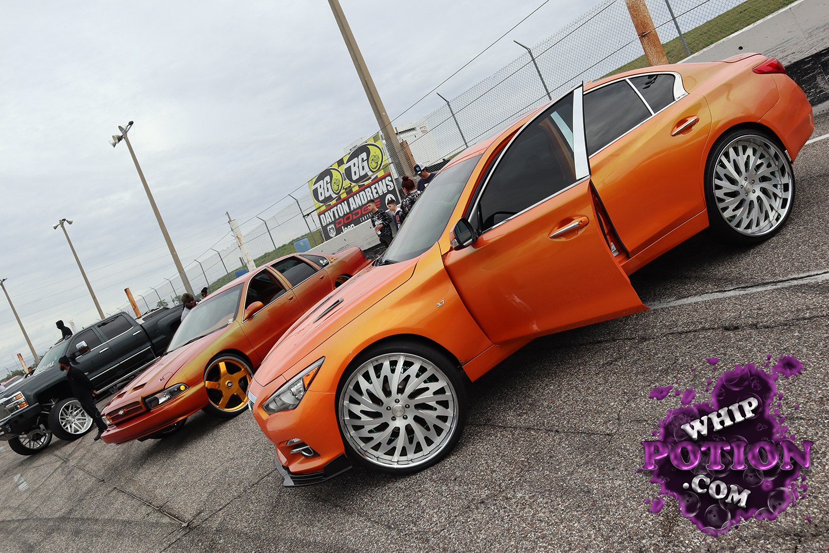 A pair of orange Infiniti sedans sits low and clean, with the foreground car wearing big chrome multi-spoke wheels that fill the fenders nicely. The rear door is open, showing a tidy custom touch rather than a full stripped-out build. Beside it, the matching sedan runs gold five-spokes and keeps the same smooth, monochrome look that gives both cars a sharp, coordinated street-show vibe.