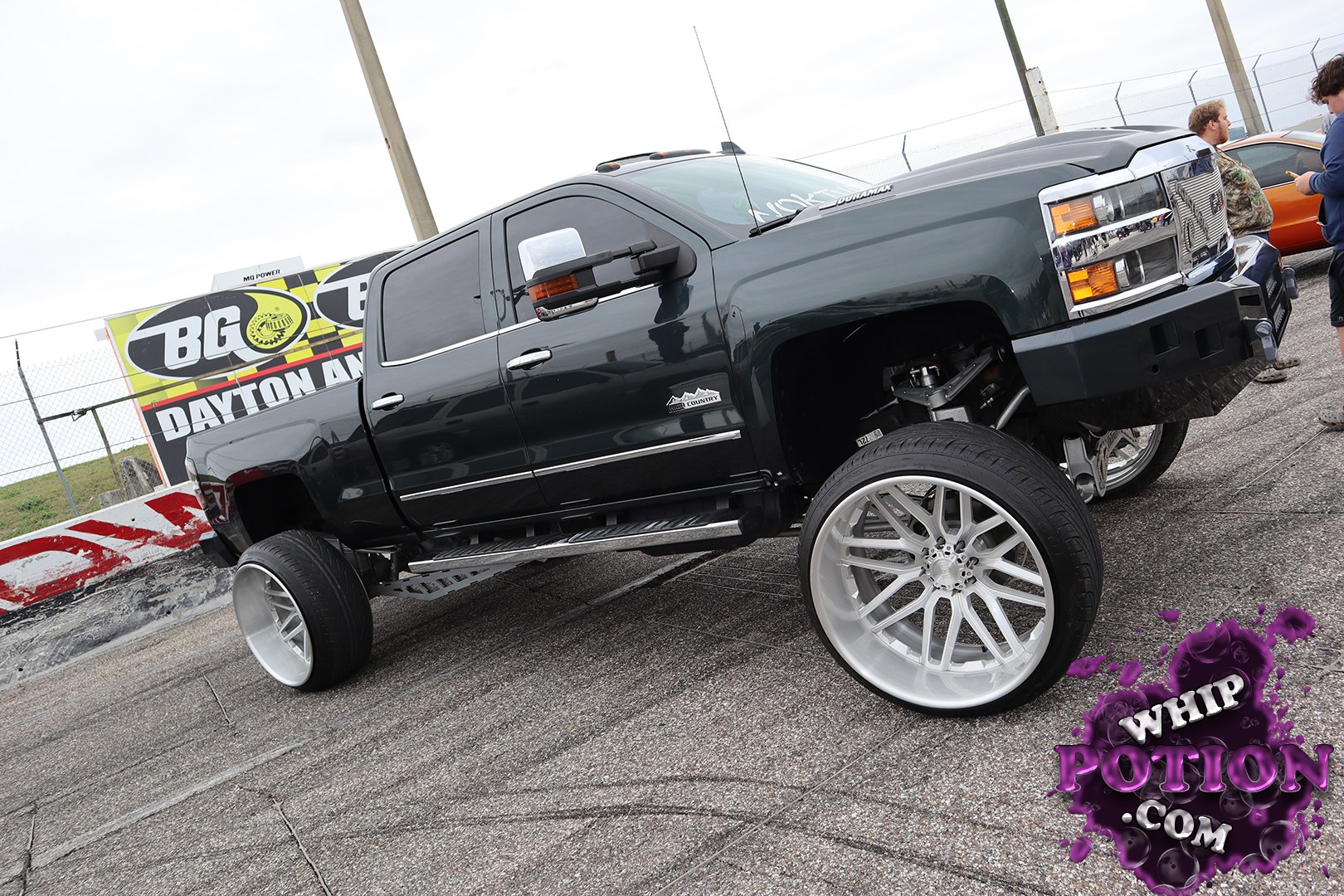 A heavy-duty Chevrolet Silverado 2500HD gets the full big-wheel treatment here, with a dark paint finish, massive polished multi-spokes, and a sky-high suspension setup that exposes plenty of custom underpinnings. The stance is unapologetically bold, and the chiseled front end with its chrome grille and tow-spec attitude gives it real street-truck presence.