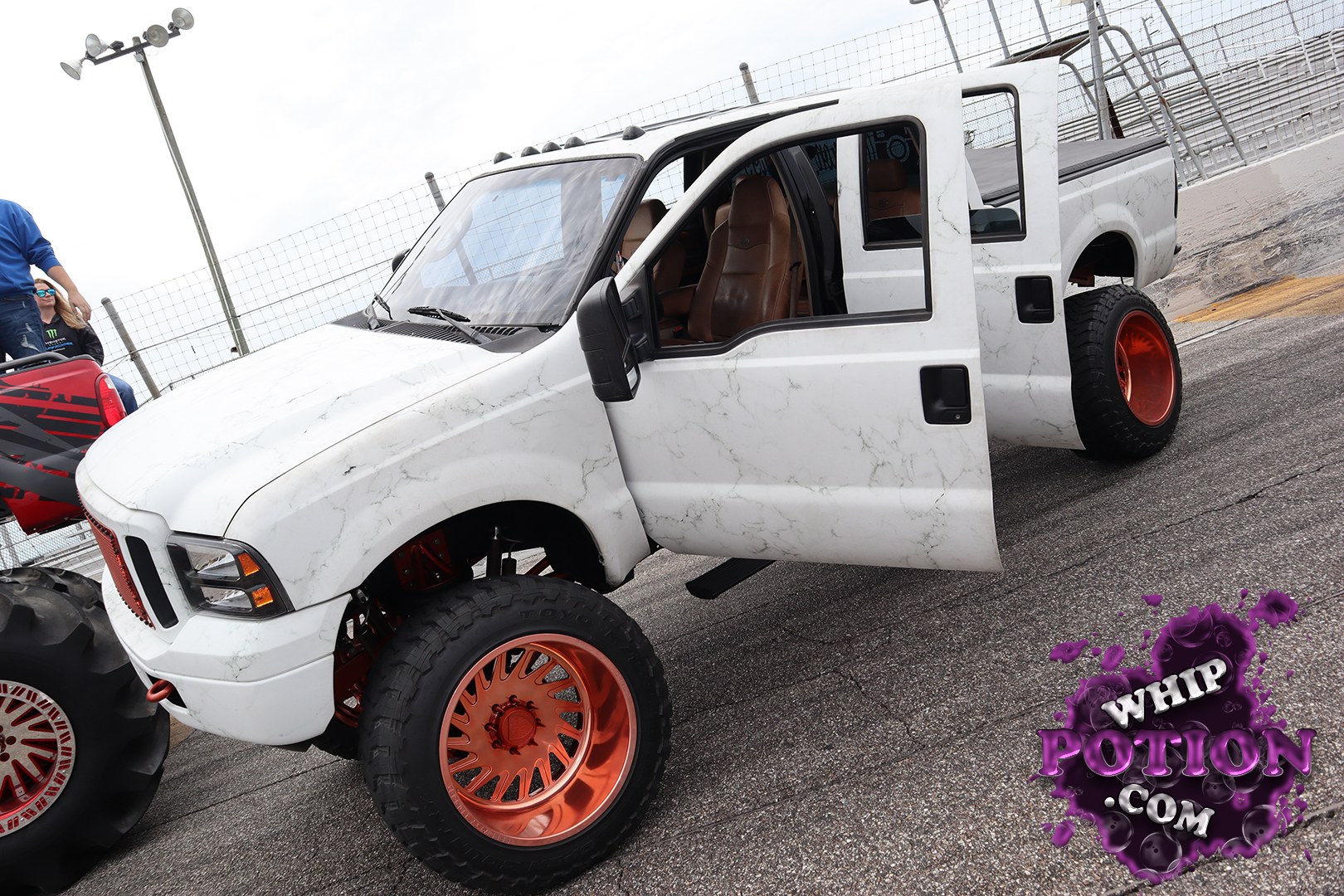 This lifted Dodge Ram crew cab leans hard into custom truck culture, with a white marble-style wrap, oversized copper wheels wrapped in chunky tires, and a tall stance that shows off the suspension. The brown leather interior and clean shaved look give it a high-end finish, while the bed cover keeps the build looking tight and purposeful.
