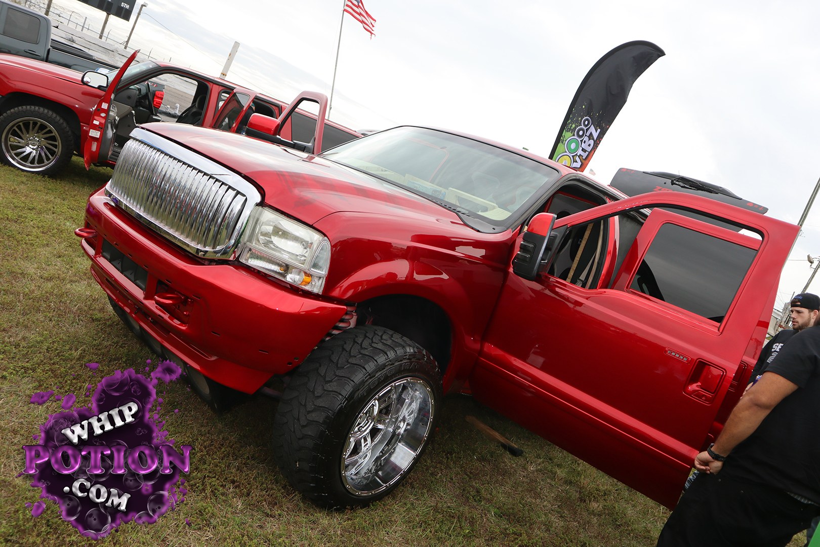 A bold red Ford Super Duty pickup sits on oversized chrome wheels with deep off-road rubber, mixing show-truck flash with serious stance. The big vertical-bar grille and square headlamps give it away, while the lowered front bumper and shaved, monochrome paint keep the build clean and aggressive. Open doors let the custom interior peek through.