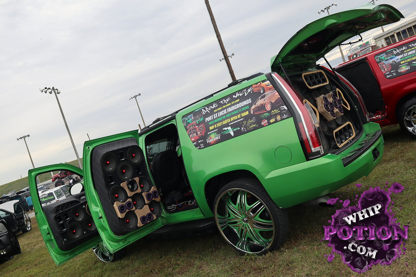 A bright green Cadillac Escalade wears a full custom audio build, with the rear hatch and both passenger-side doors opened to show off stacks of speakers, tweeters, and cleanly upholstered panels. The big billet-style green-and-chrome wheels and slammed stance give it plenty of show-car attitude, while the window decal and matching trim tie the whole setup together.