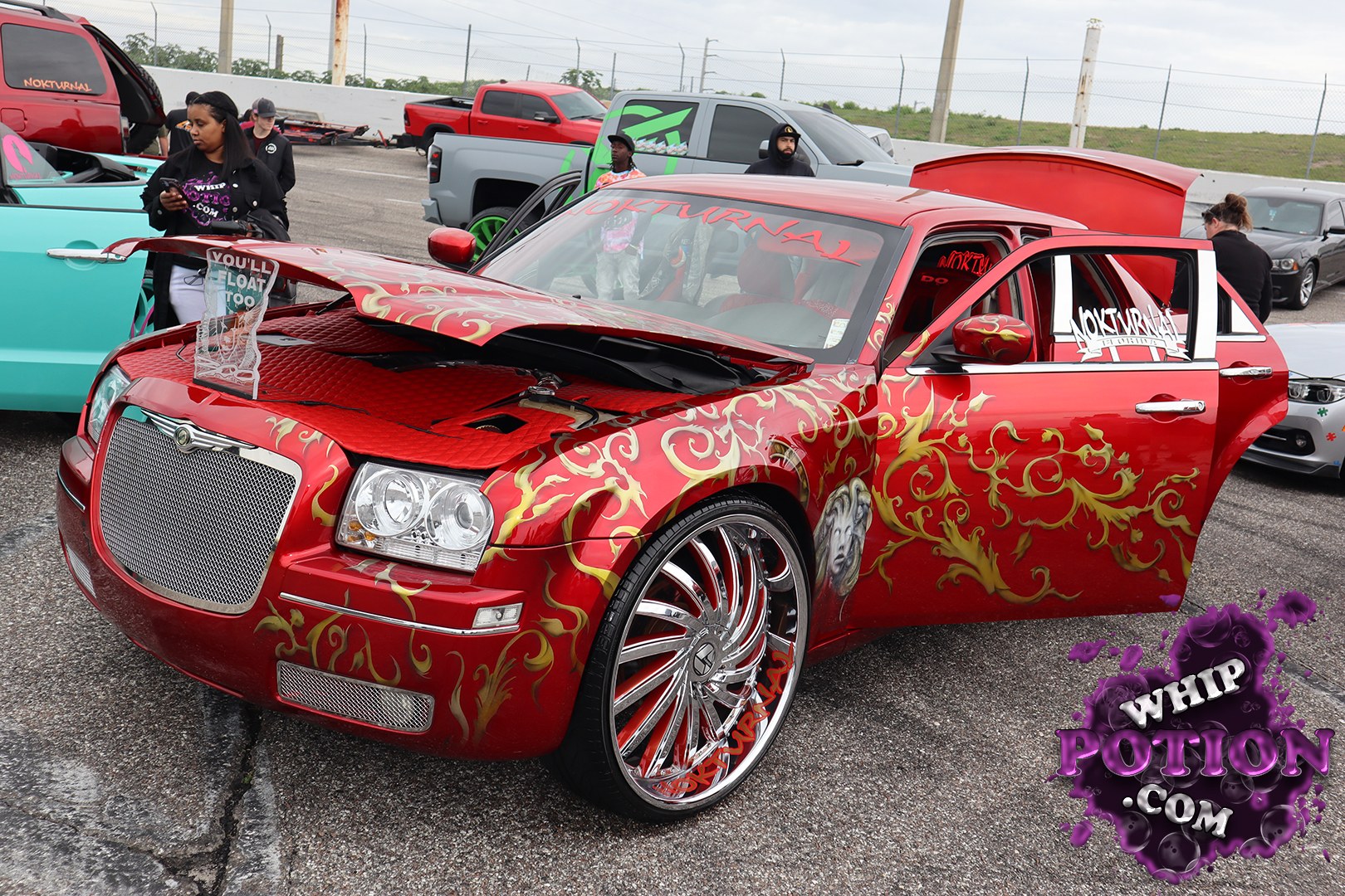 This Chrysler 300 goes full custom with a deep red candy finish, gold flame graphics, and a wild mural down the side. The hood is propped to show off the clean engine bay treatment, while the big chrome multi-spokes and low stance give it real show-car presence. The “NOKTURNAL” lettering ties the whole build together with some serious street flair.