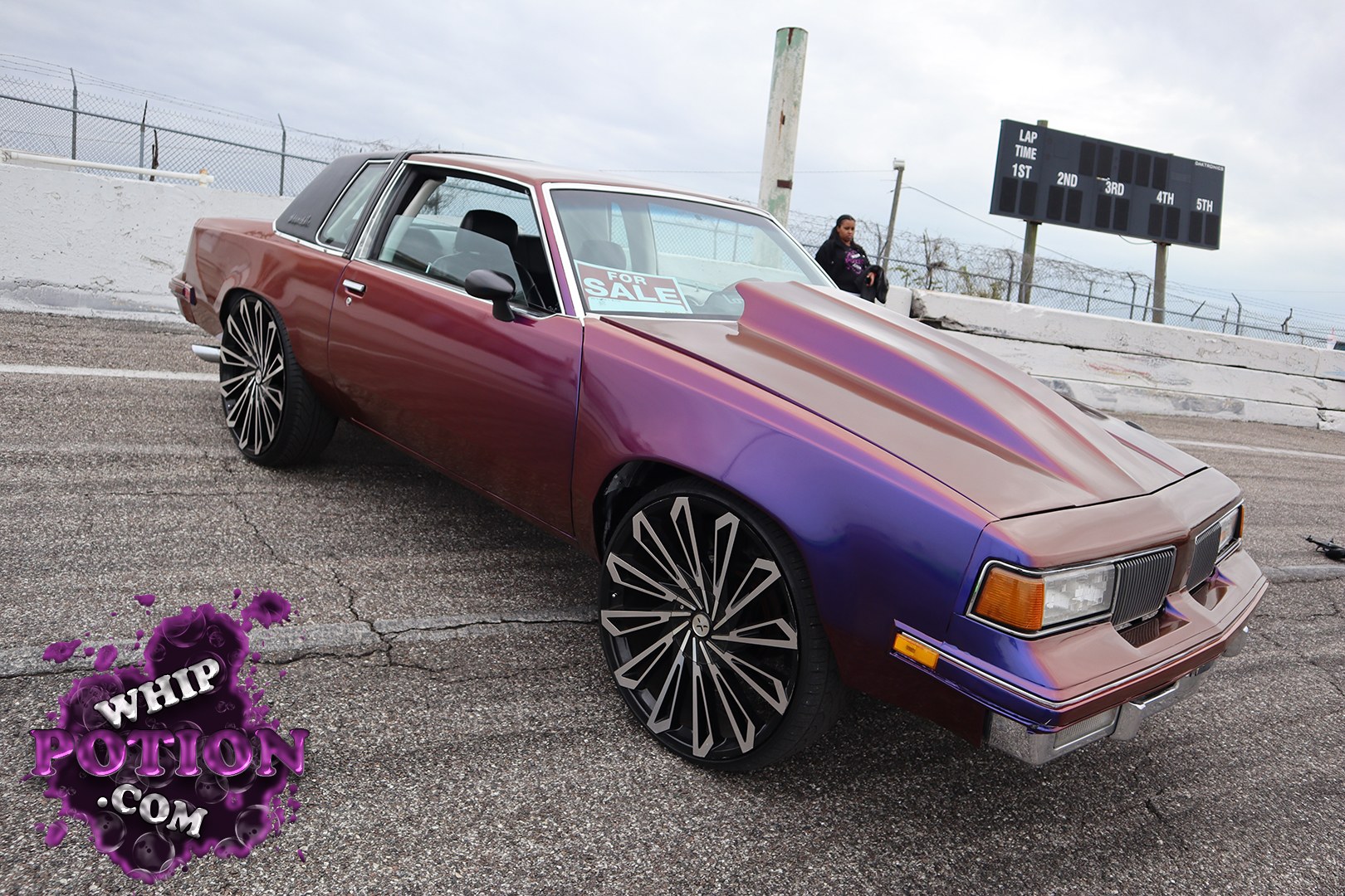 A classic coupe wears a deep chameleon fade from burgundy to violet, with a black vinyl top and a long, sharpened hood that gives it real street presence. The big multi-spoke wheels fill the wells nicely, and the clean chrome trim, tinted glass, and crisp body lines keep the whole build looking polished and intentional.