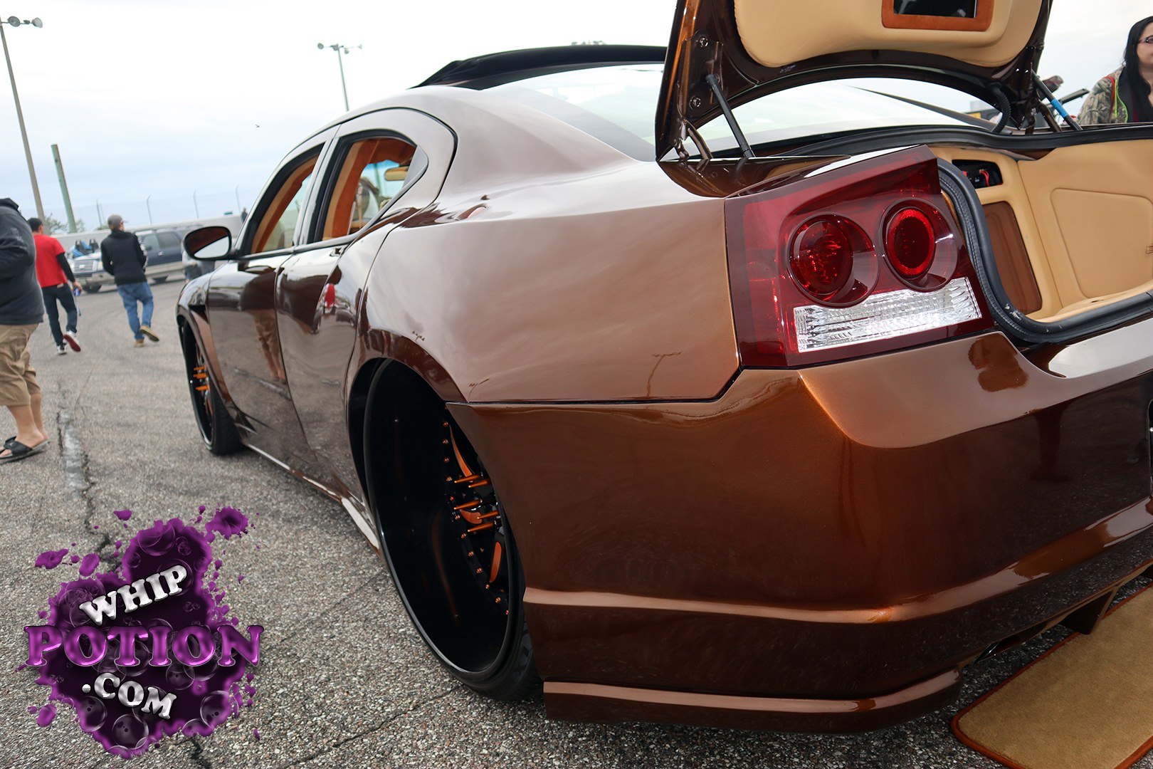 This Dodge Charger sits low and mean in a deep brown metallic finish, with oversized black wheels, orange suspension accents, and a tan custom interior visible through the open trunk and windows. The smooth bodywork and tucked stance give it a clean show-car feel, and the setup at Showtime Speedway for Linny J Official MLK Weekend Car Show 2021 looks dialed in for the loud end of the hobby.
