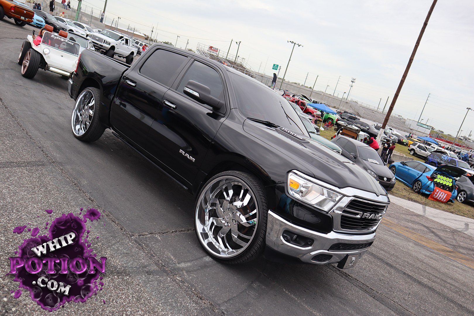 A black Ram 1500 lays out on oversized chrome billets, and the clean monochrome treatment gives it a sharp show-truck feel. The tinted cab, smooth paint, and aggressive stance keep the focus on those huge wheels tucked under the arches, while the little wild roadster in the background adds a nice slice of car-culture chaos.
