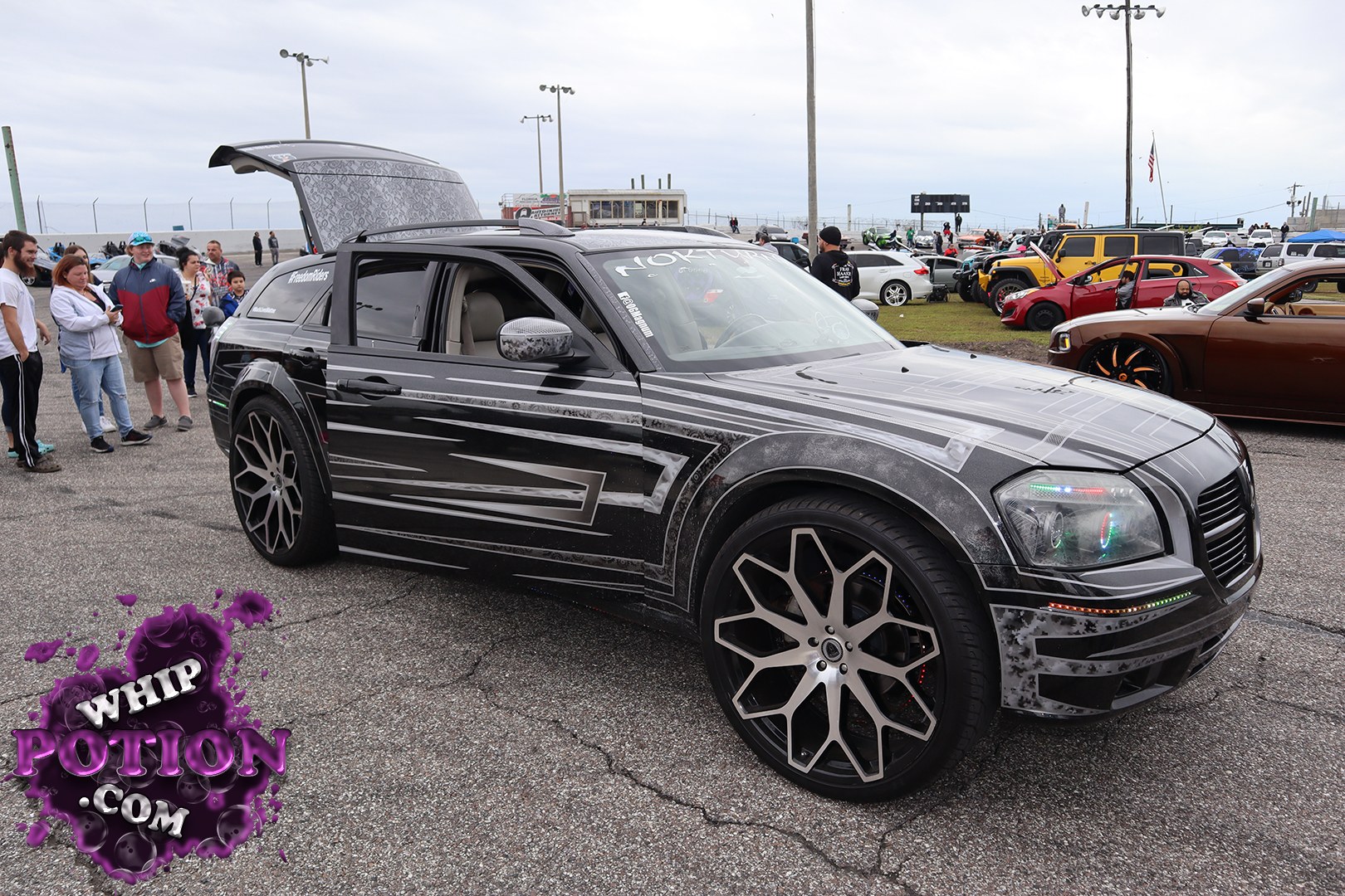 A Chrysler 300 wagon conversion with a wild custom wrap, suicide-style rear hatch display, and oversized multi-spoke wheels gives this build a full VIP-meets-show-car vibe. The black-and-gray graphics flow cleanly over the body, while the tinted glass, tucked trim, and clean stance make it look ready for the next round of judging.