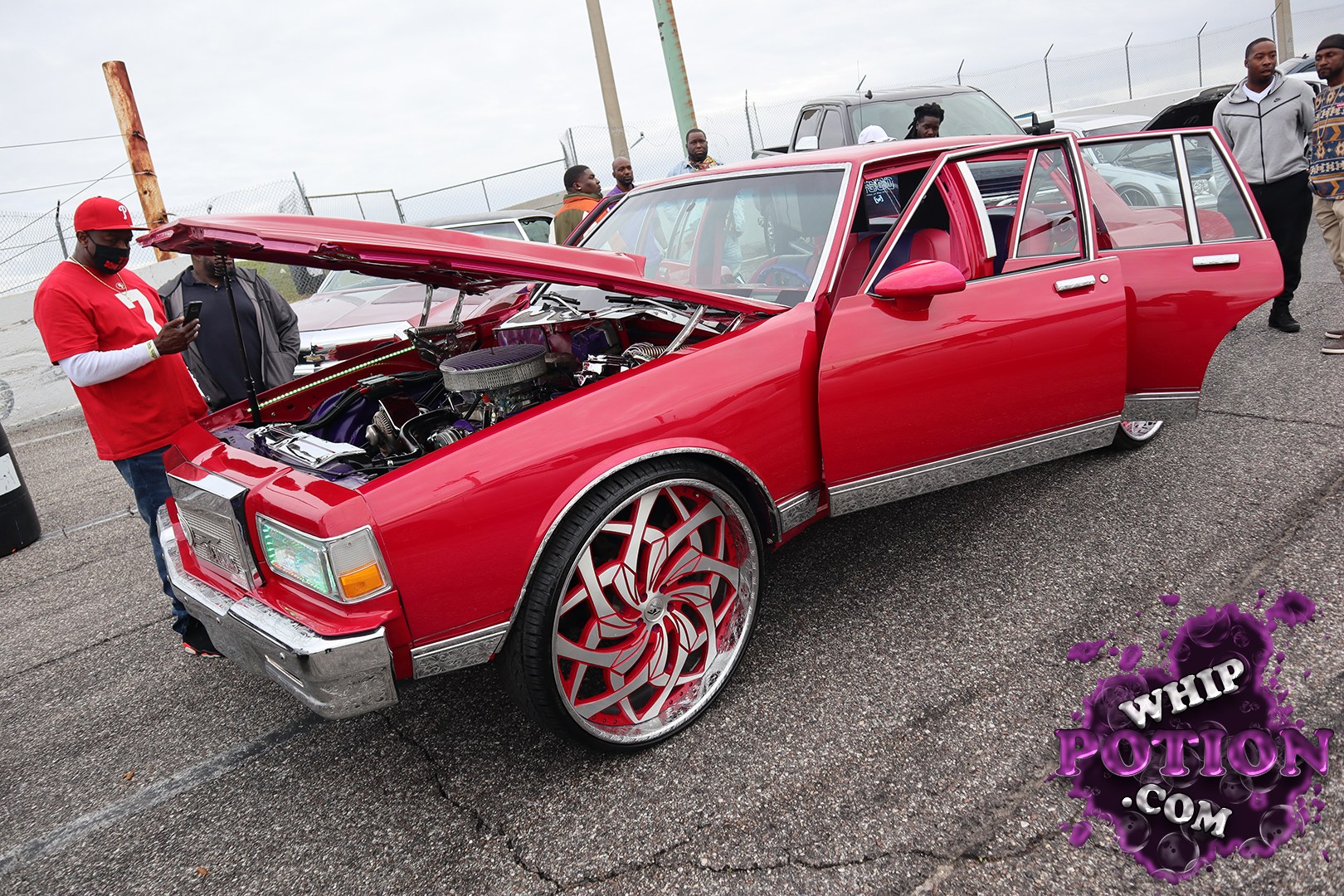 This boxy GMC wagon has been fully personalized in bright red, with the hood propped open to show off a cleaned-up, detailed engine bay and a custom-cabin look inside. The oversized red-and-chrome billet wheels, chrome trim, and polished bumper give it a bold, street-show attitude, while the lowered stance keeps the whole build sitting just right.