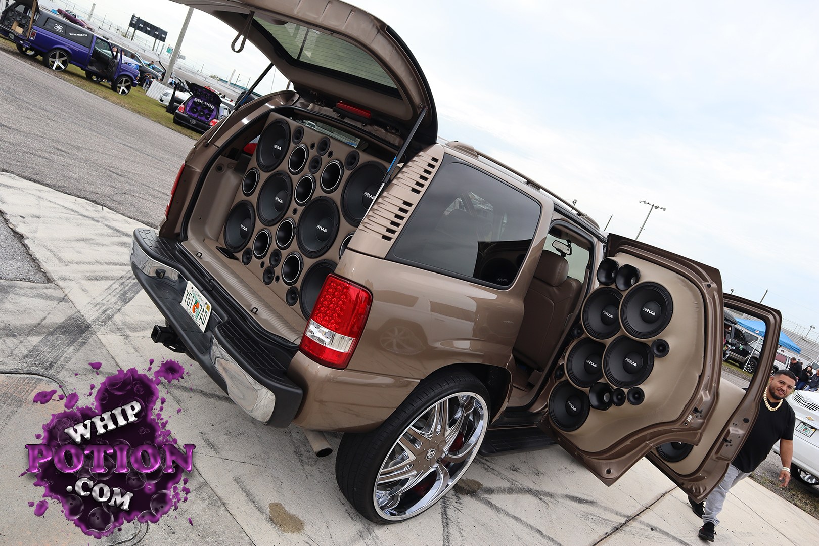 This full-size SUV is built for show and sound, with both rear doors and the tailgate open to reveal a wall of PRV speakers packed into custom tan door panels and cargo trim. The bronze paint, massive chrome wheels, low stance, and tinted glass give it a clean, hard-hitting look that’s all about presence and serious audio swagger.
